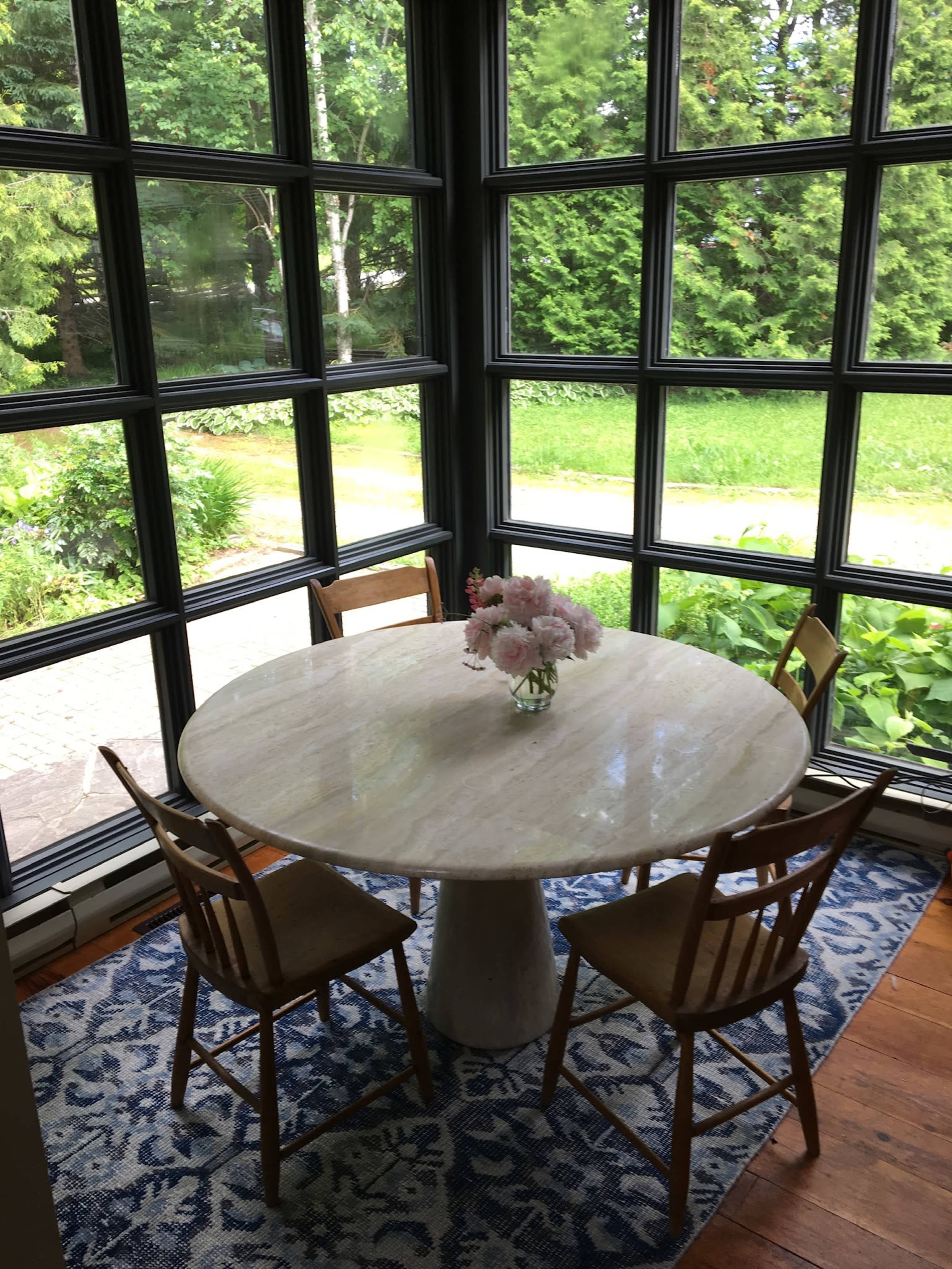 A round marble table with four wooden chairs is set on a blue patterned rug in a sunlit room surrounded by large windows overlooking a green garden.