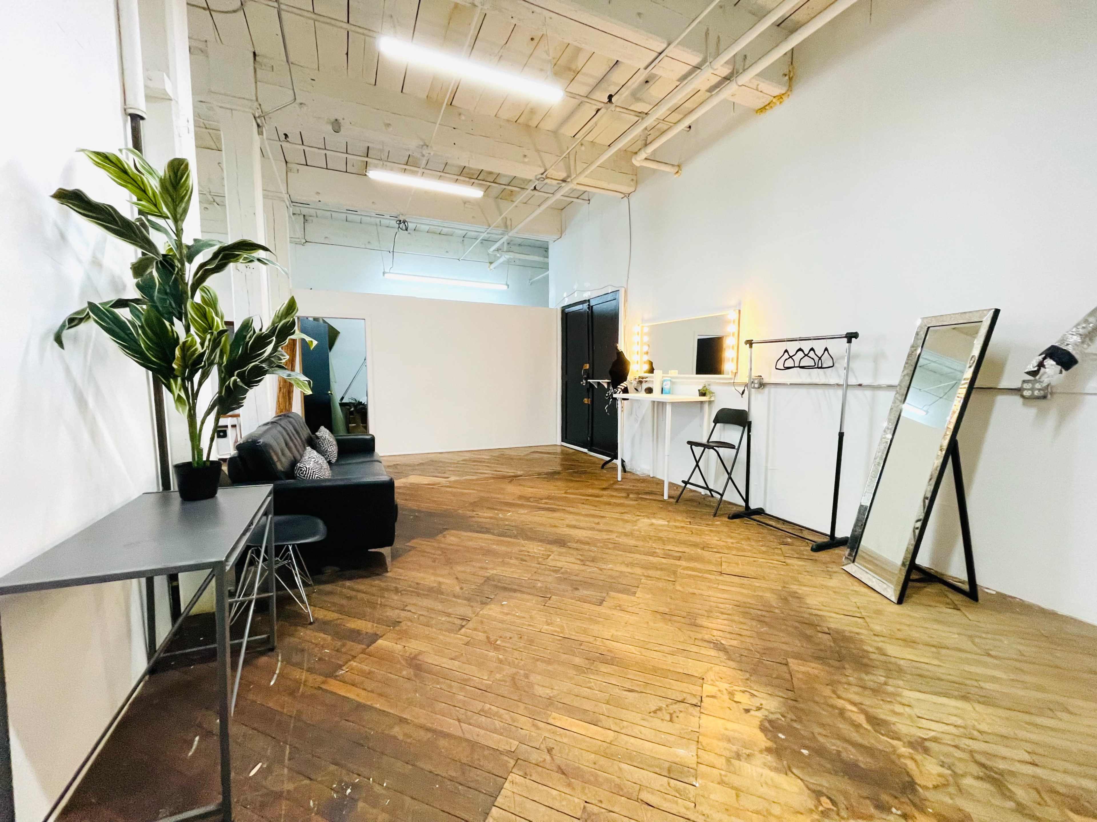 A spacious interior room features wooden flooring, a couch, a tall mirror, a table with a chair, and a wall-mounted vanity with lights.