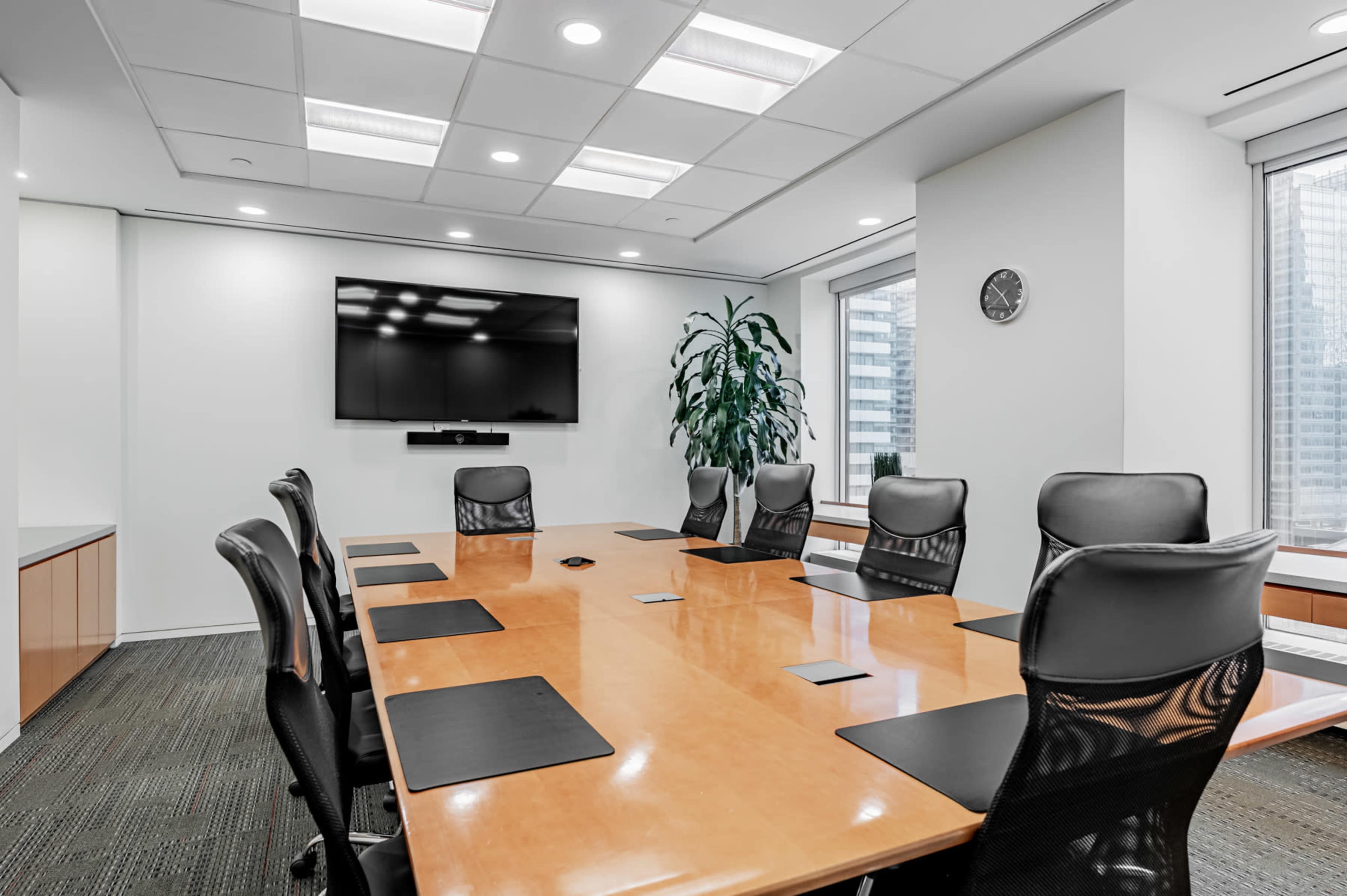 A modern conference room features a large wooden table surrounded by ergonomic black chairs, with a wall-mounted TV and a clock visible.
