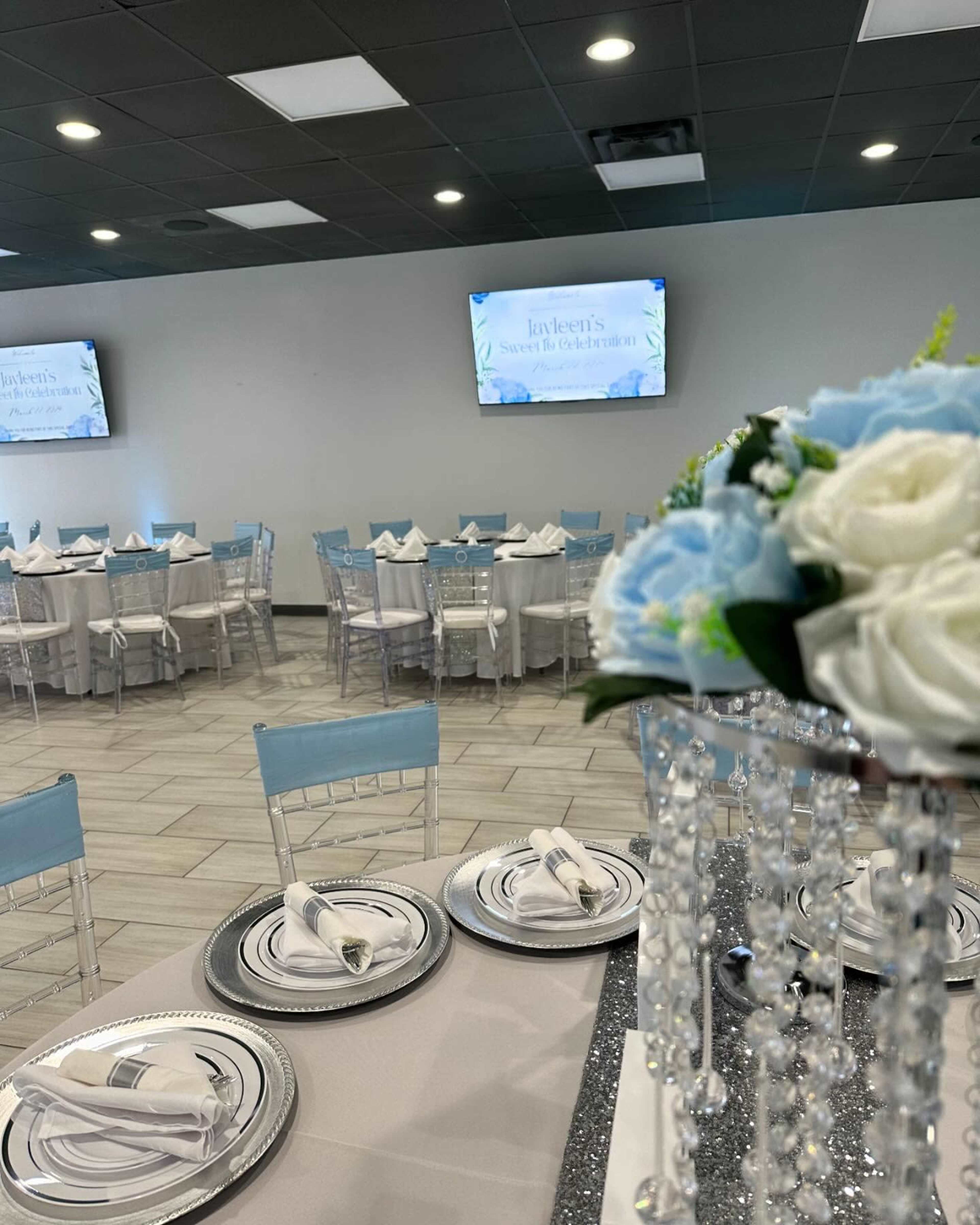 The image shows a banquet hall set up for an event, featuring tables elegantly arranged with decorative linens, centerpieces, and screens displaying a presentation.