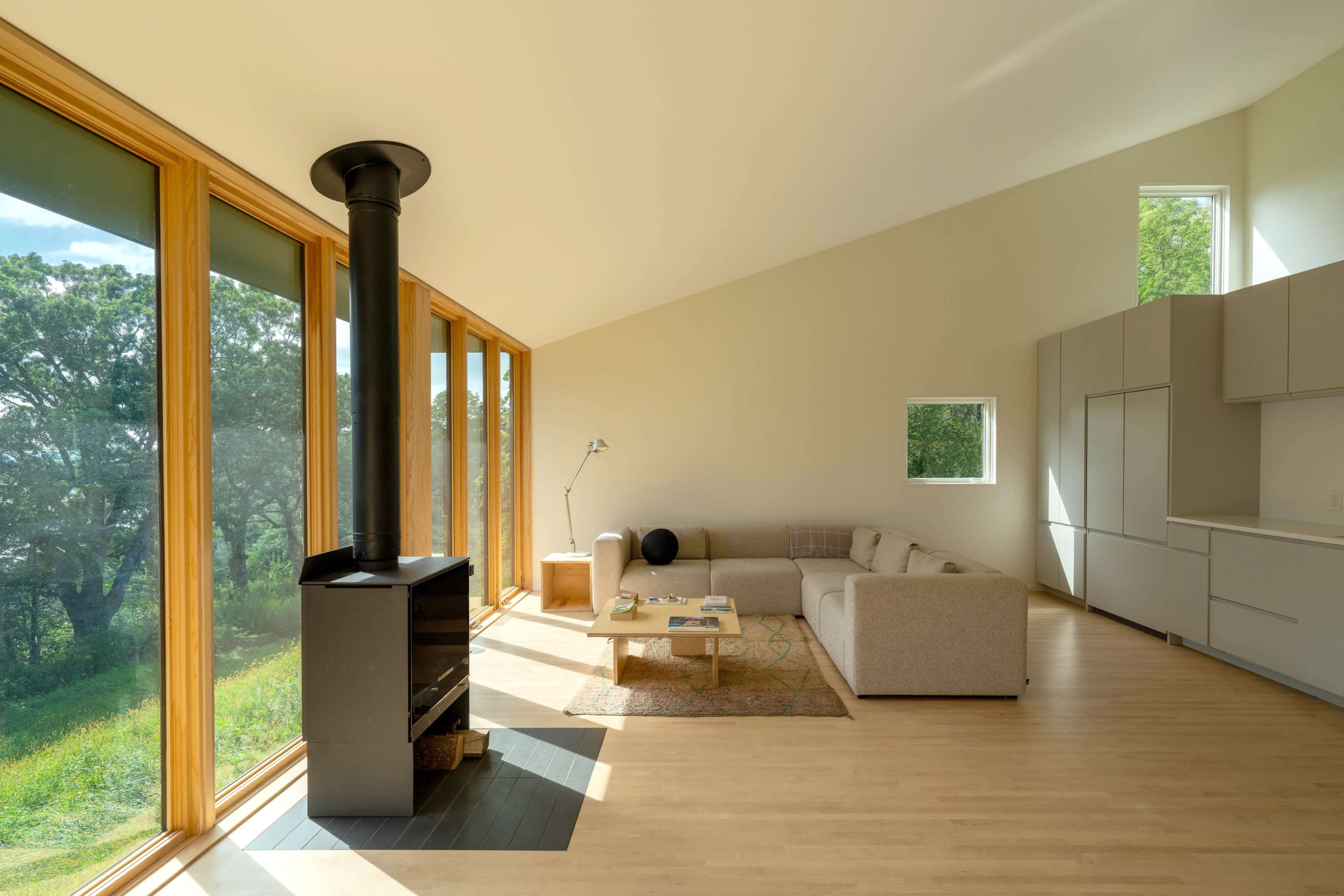 A modern living room features large windows, a light-colored sectional sofa, a wood stove, and minimalist furniture against a backdrop of greenery.