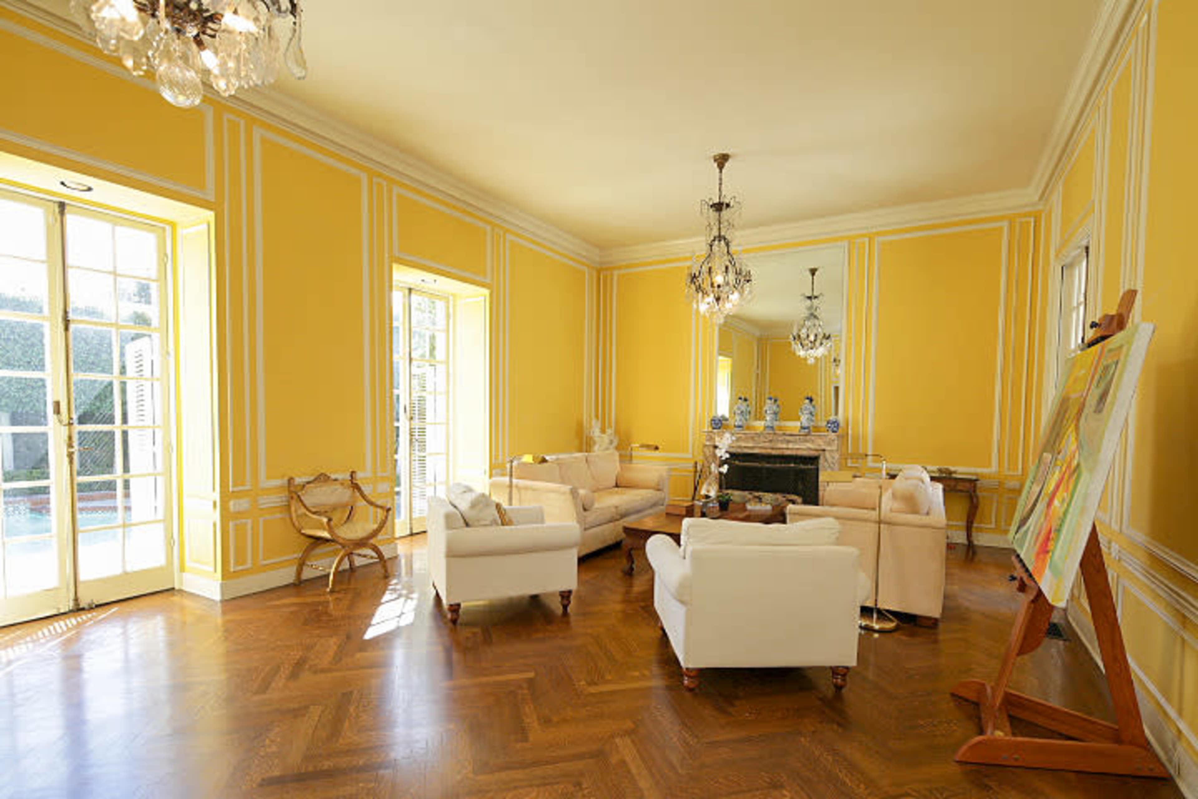 The room features yellow walls, a hardwood floor, and several seating arrangements centered around a coffee table, with large windows allowing natural light.