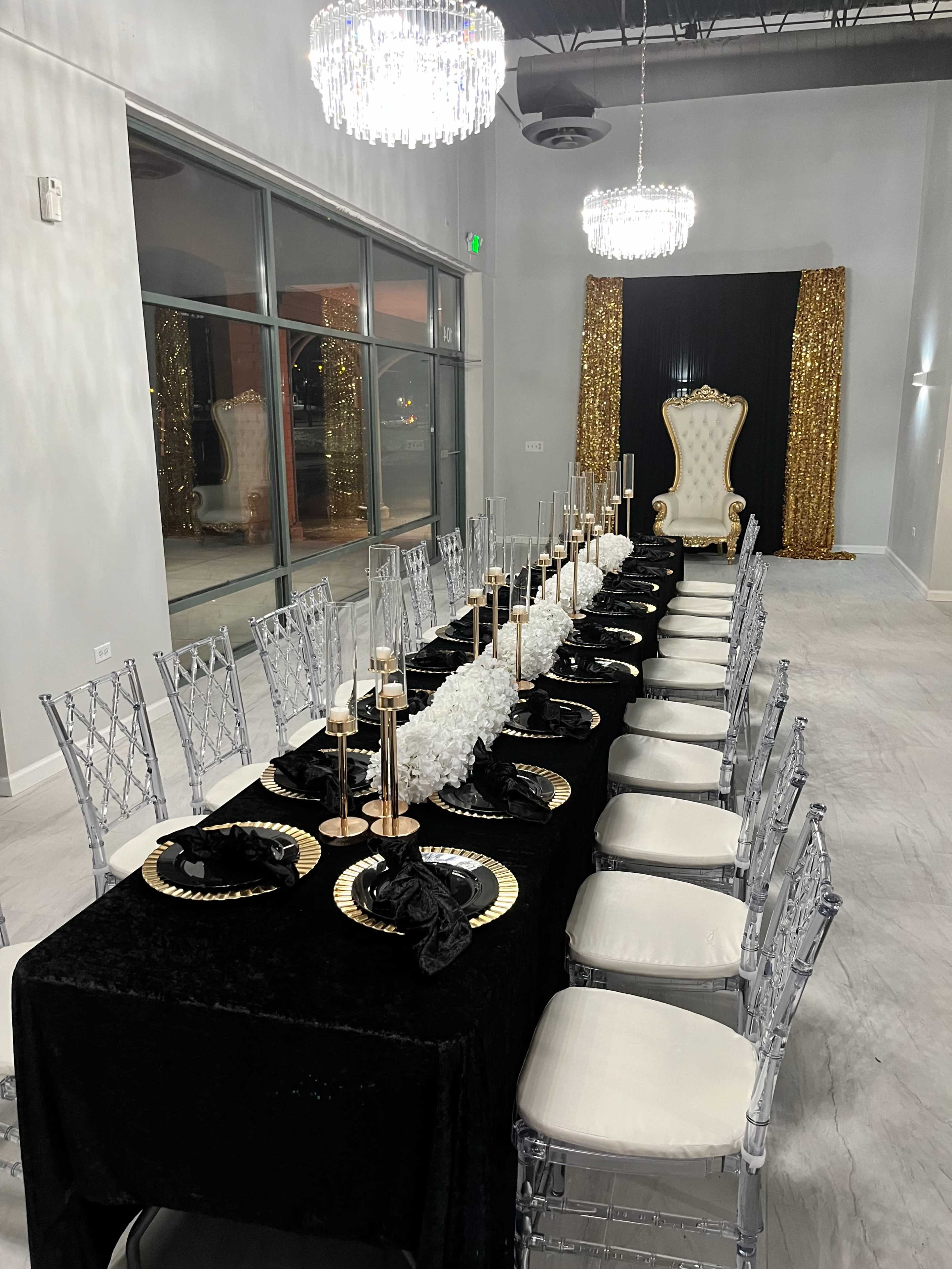 The image shows an elegantly set dining table with black and gold decor, surrounded by clear acrylic chairs in a well-lit indoor venue.
