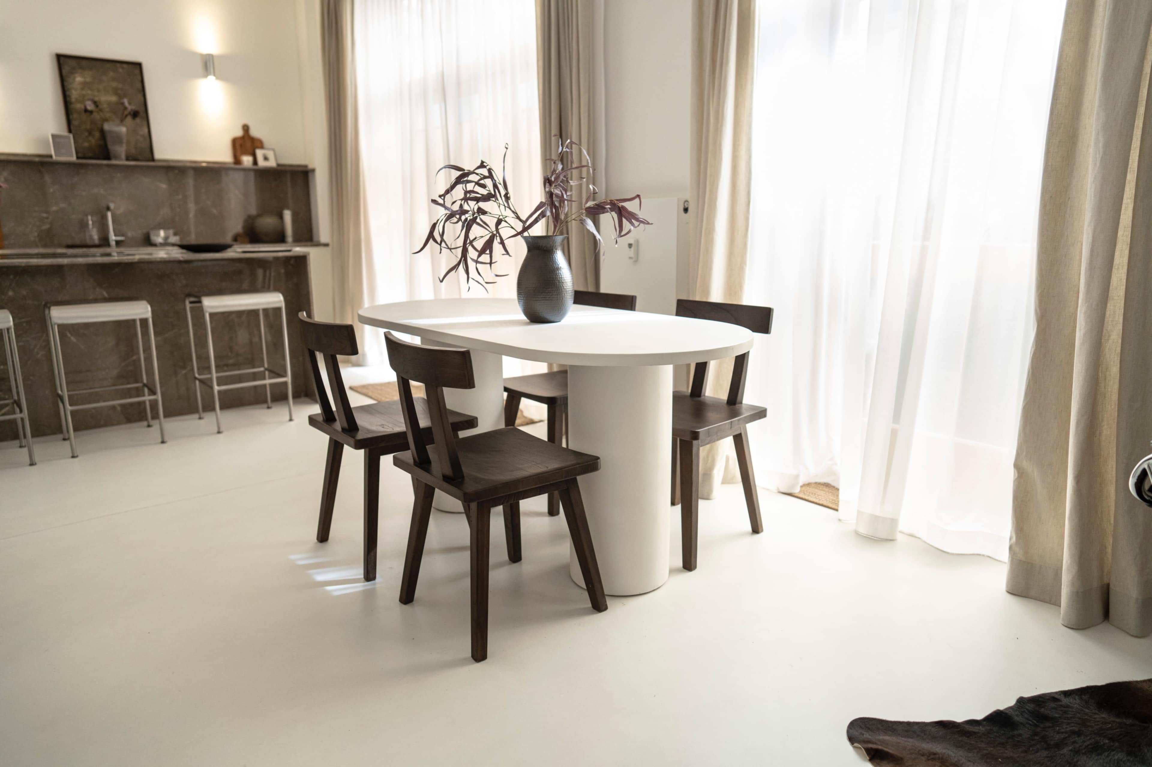 A round white dining table surrounded by four wooden chairs is positioned in a bright room with large windows and light curtains.