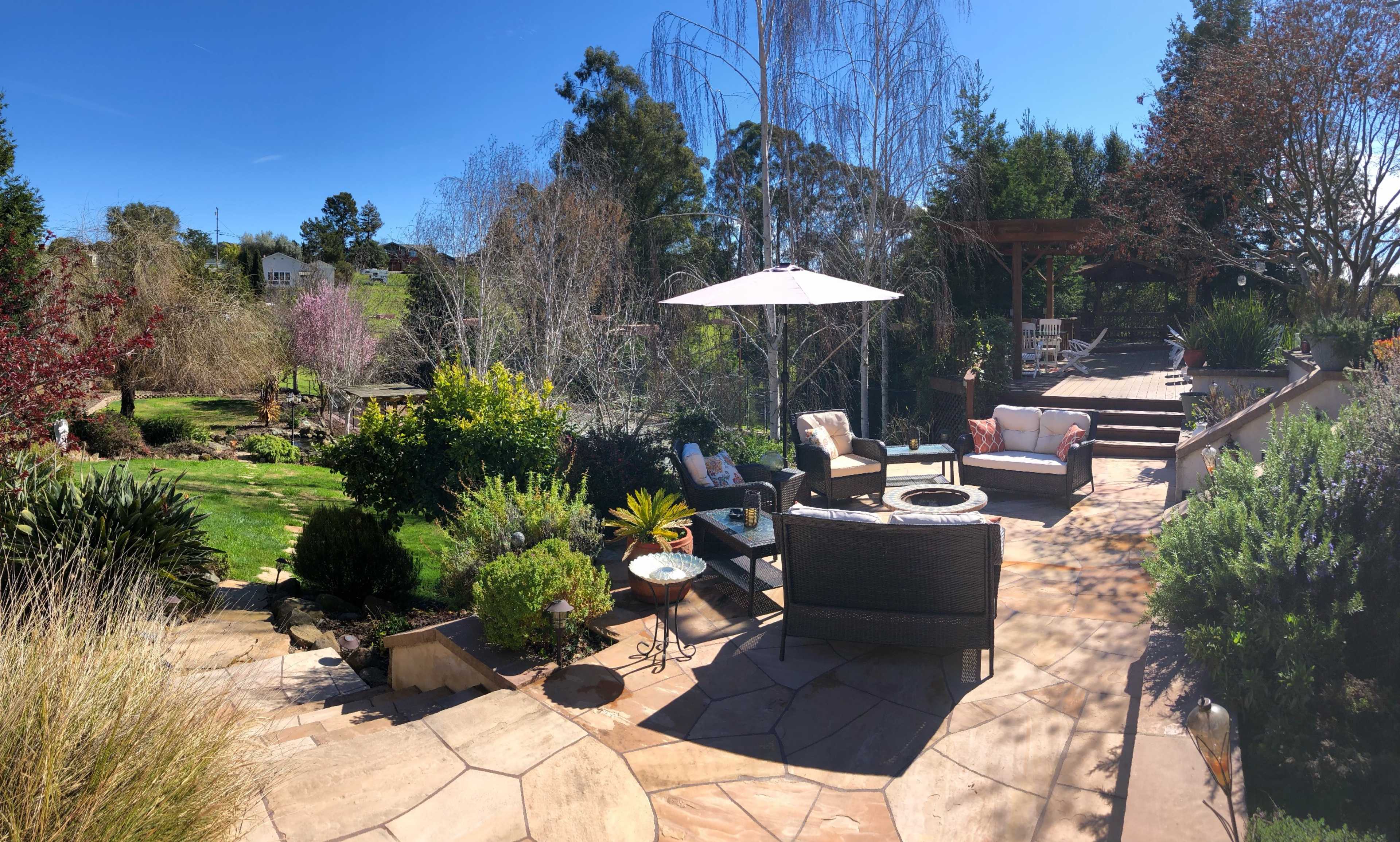 A landscaped outdoor patio area with seating arrangements, surrounding greenery, and a patio umbrella under a clear blue sky.