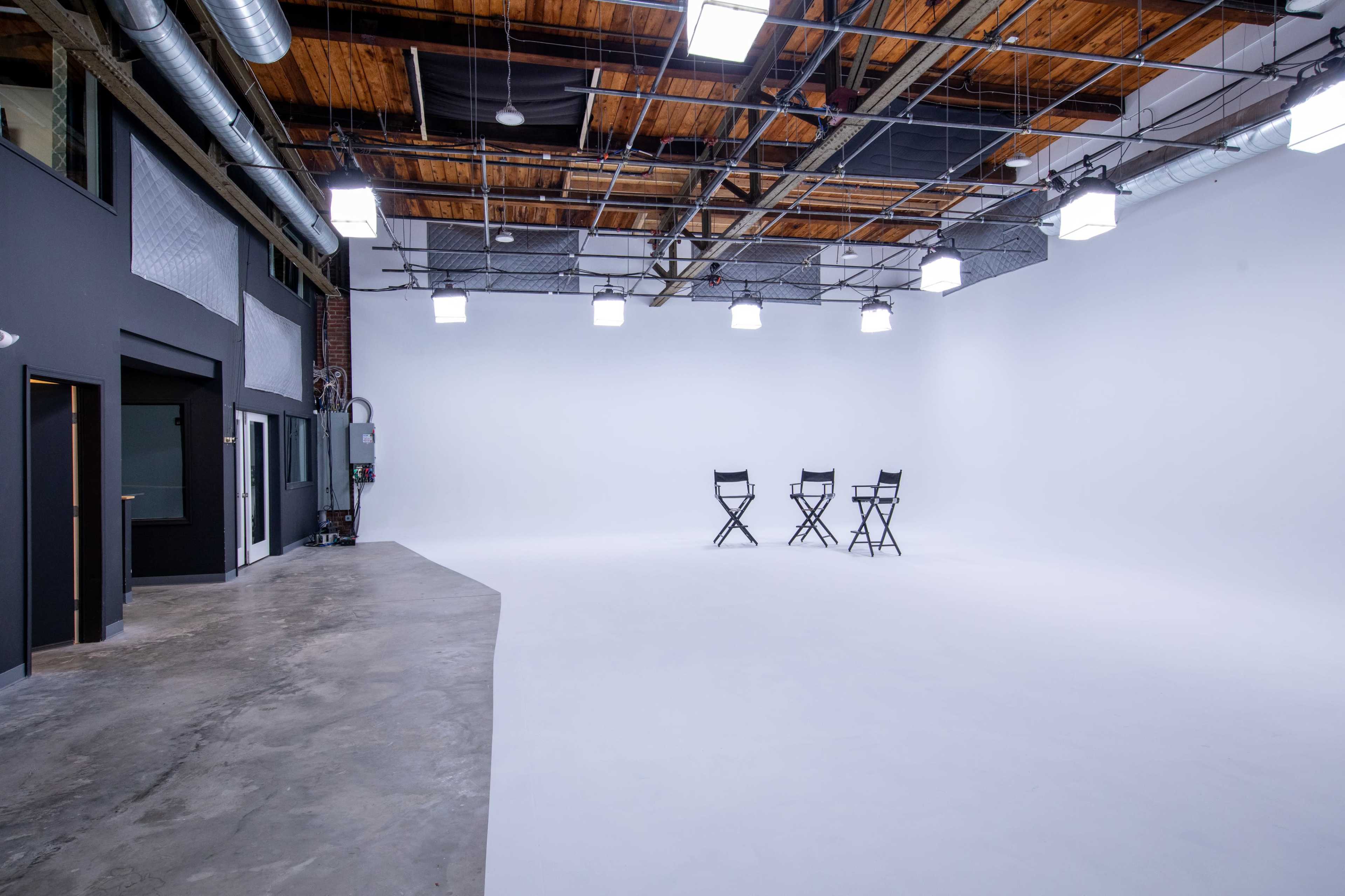 A spacious studio with a clean white backdrop, three director's chairs, and overhead lighting fixtures.