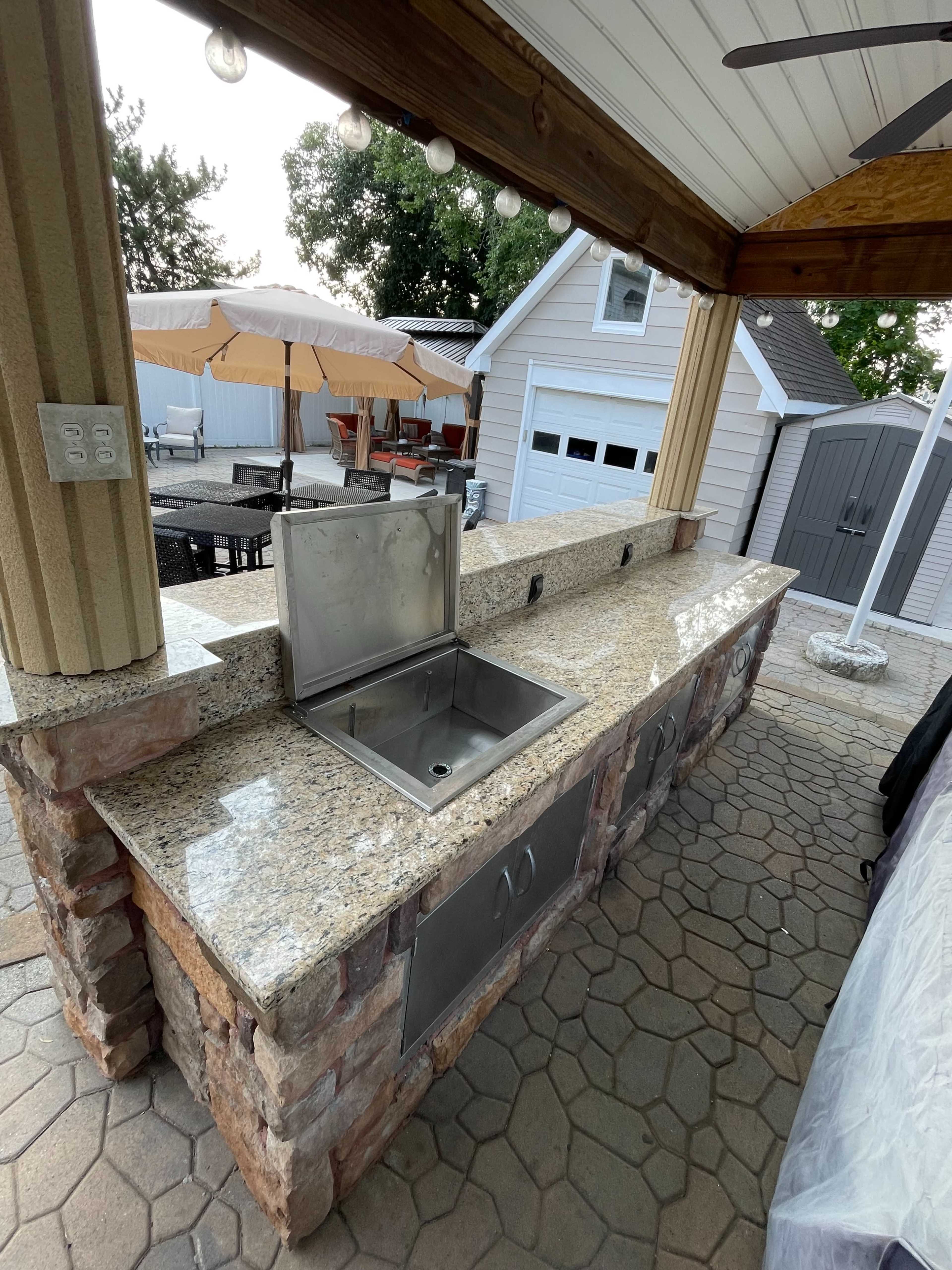 The image shows an outdoor kitchen area featuring a stone and granite countertop with a stainless steel sink and storage space, surrounded by a patio.