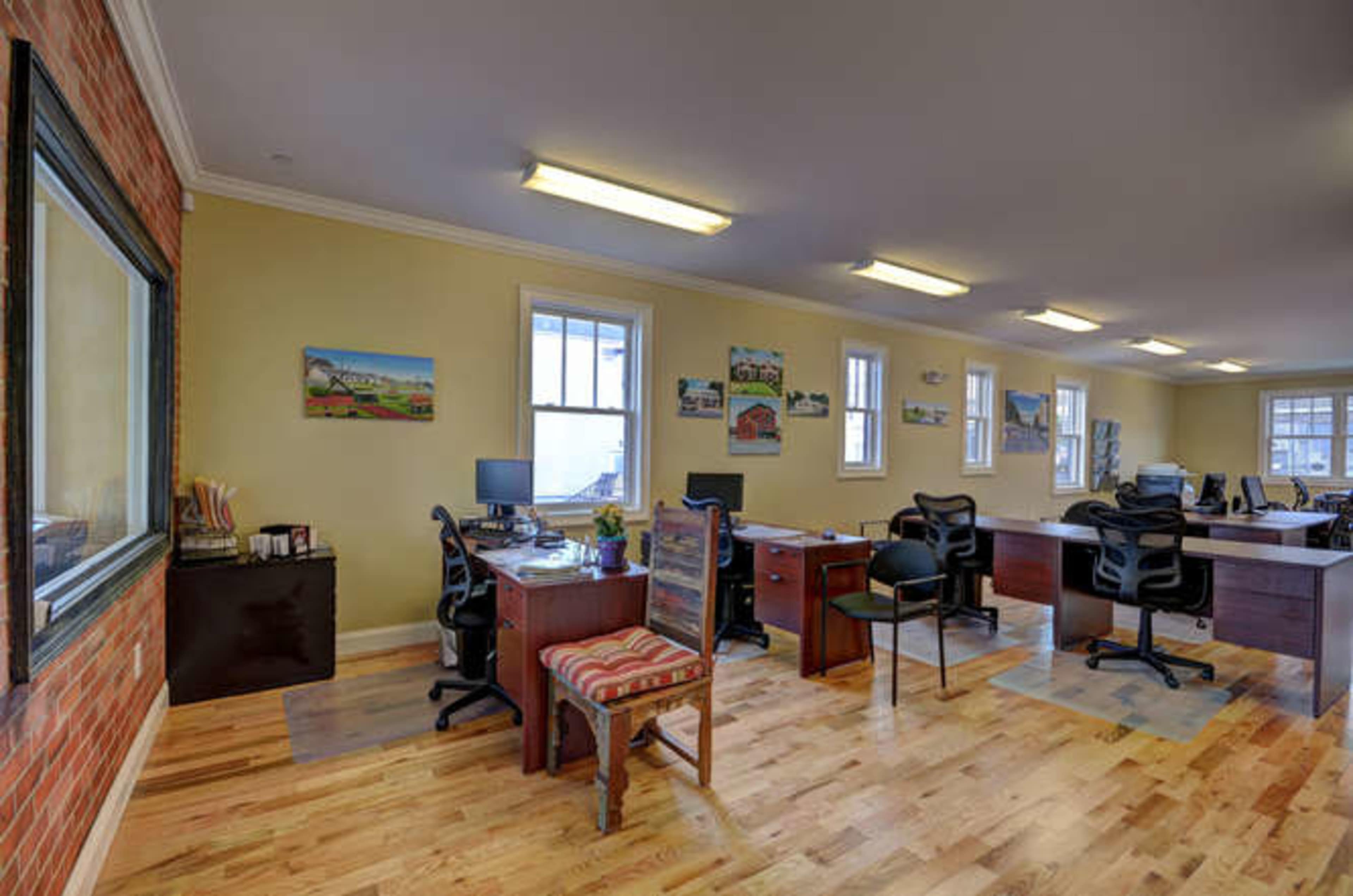 The image shows an office space featuring multiple desks, chairs, and artwork on the walls, with a wooden floor and large windows allowing natural light.