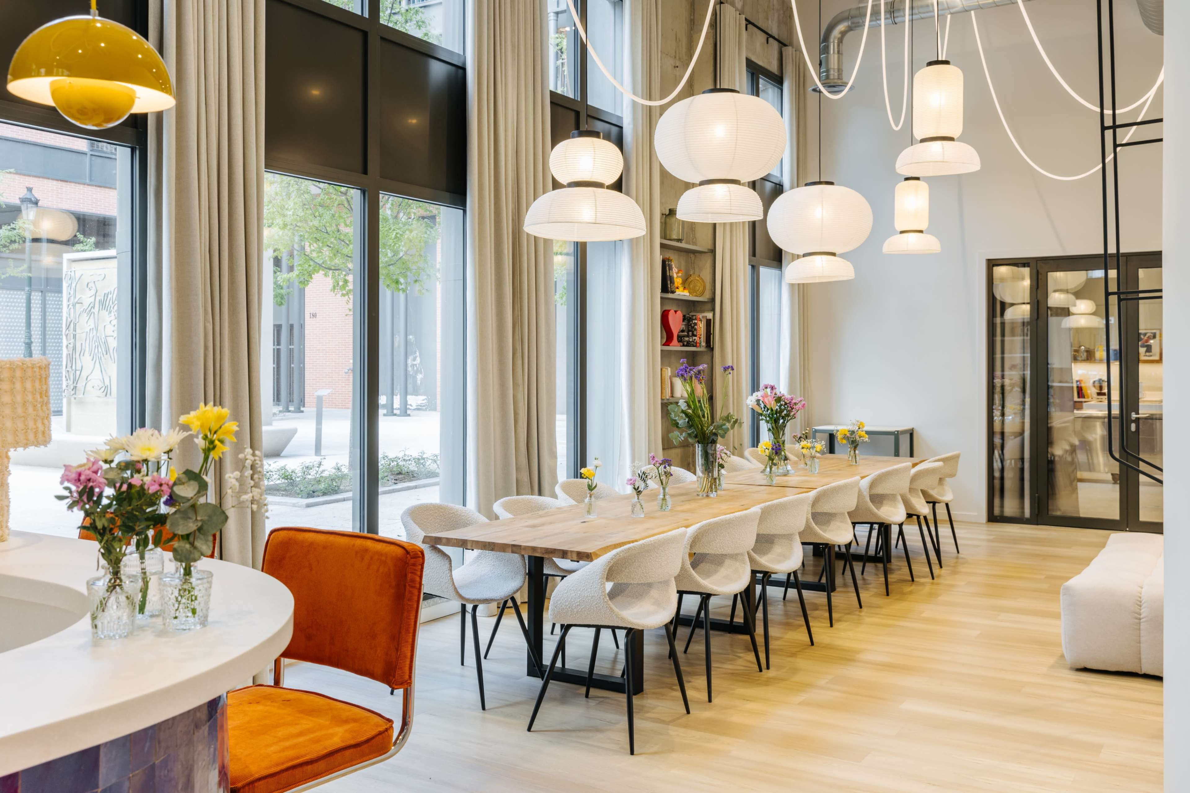 The image shows a modern dining area with a long wooden table, surrounded by white chairs, and illuminated by multiple pendant lights.