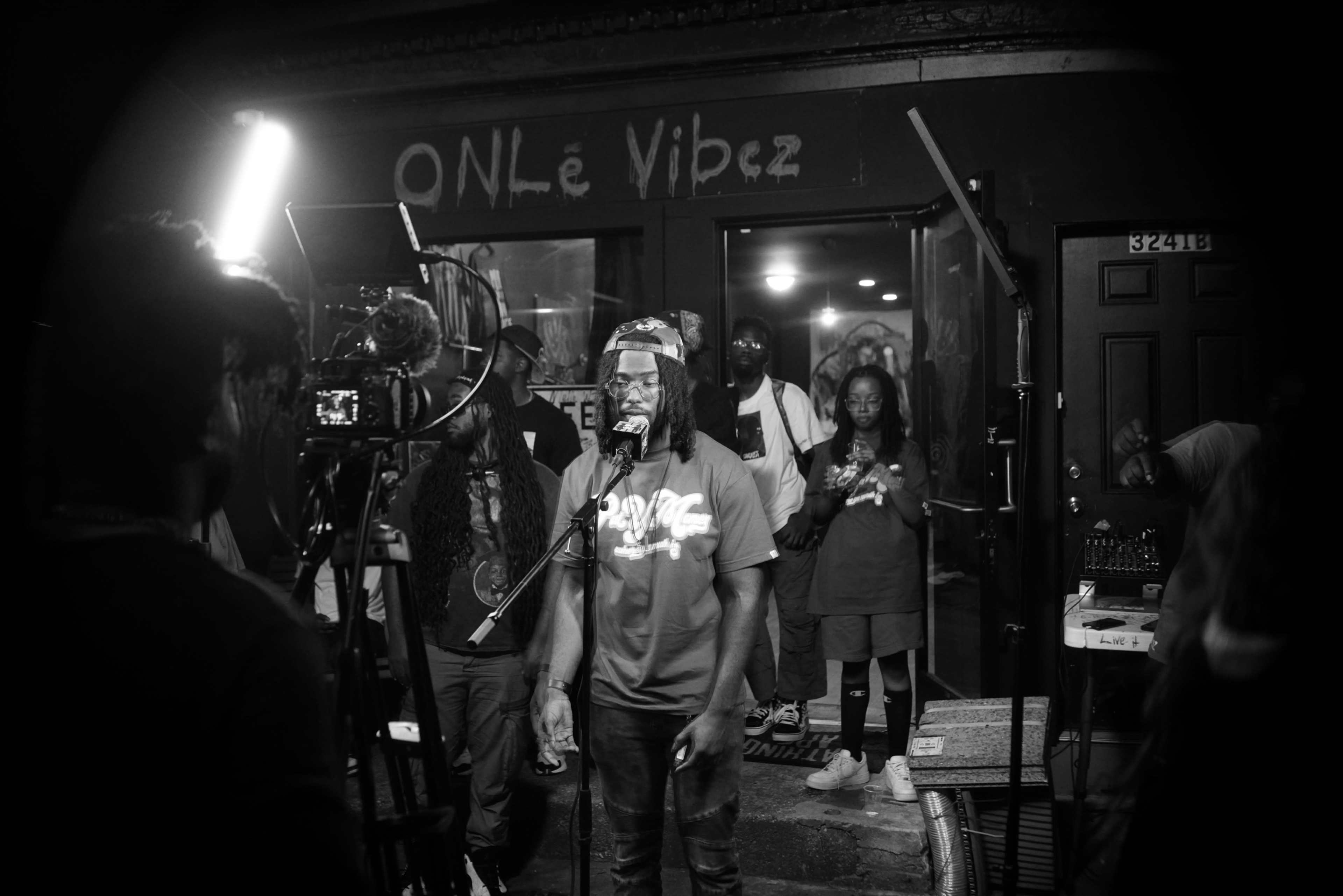 A group of performers and audience members gathers outside a venue called "ONLe Vibez," where one musician is speaking into a microphone.