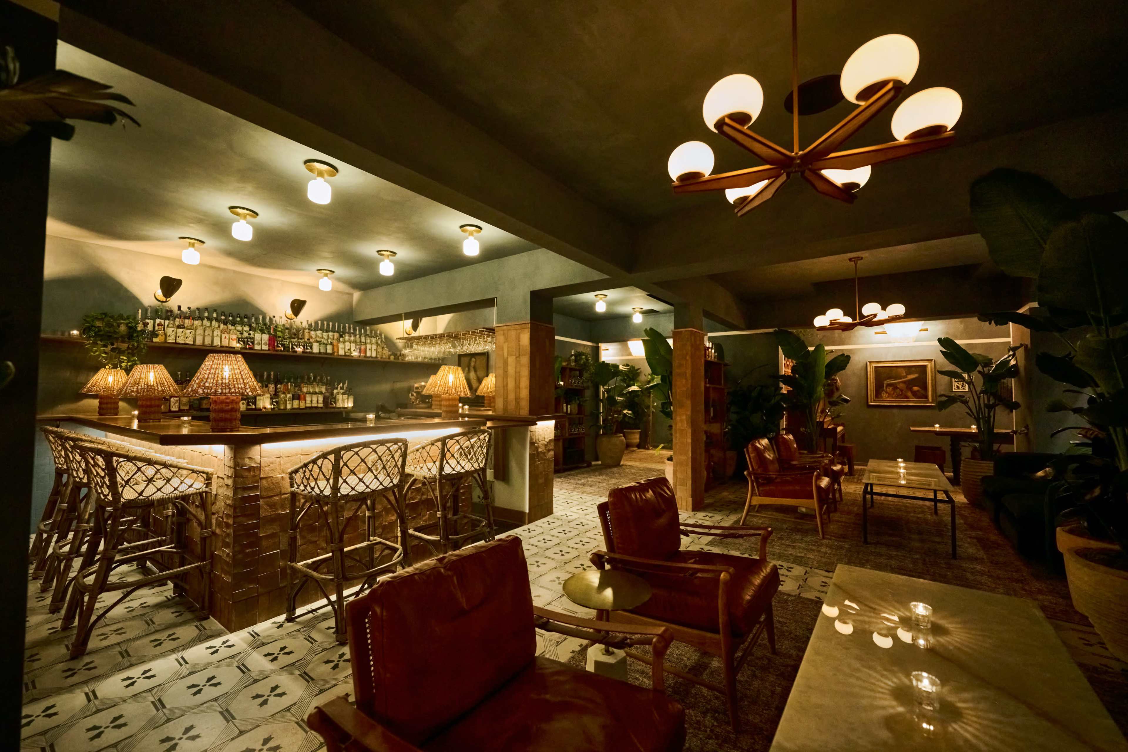 A well-lit bar with a wooden counter and high chairs is flanked by cozy seating areas adorned with plants and decorative lamps.