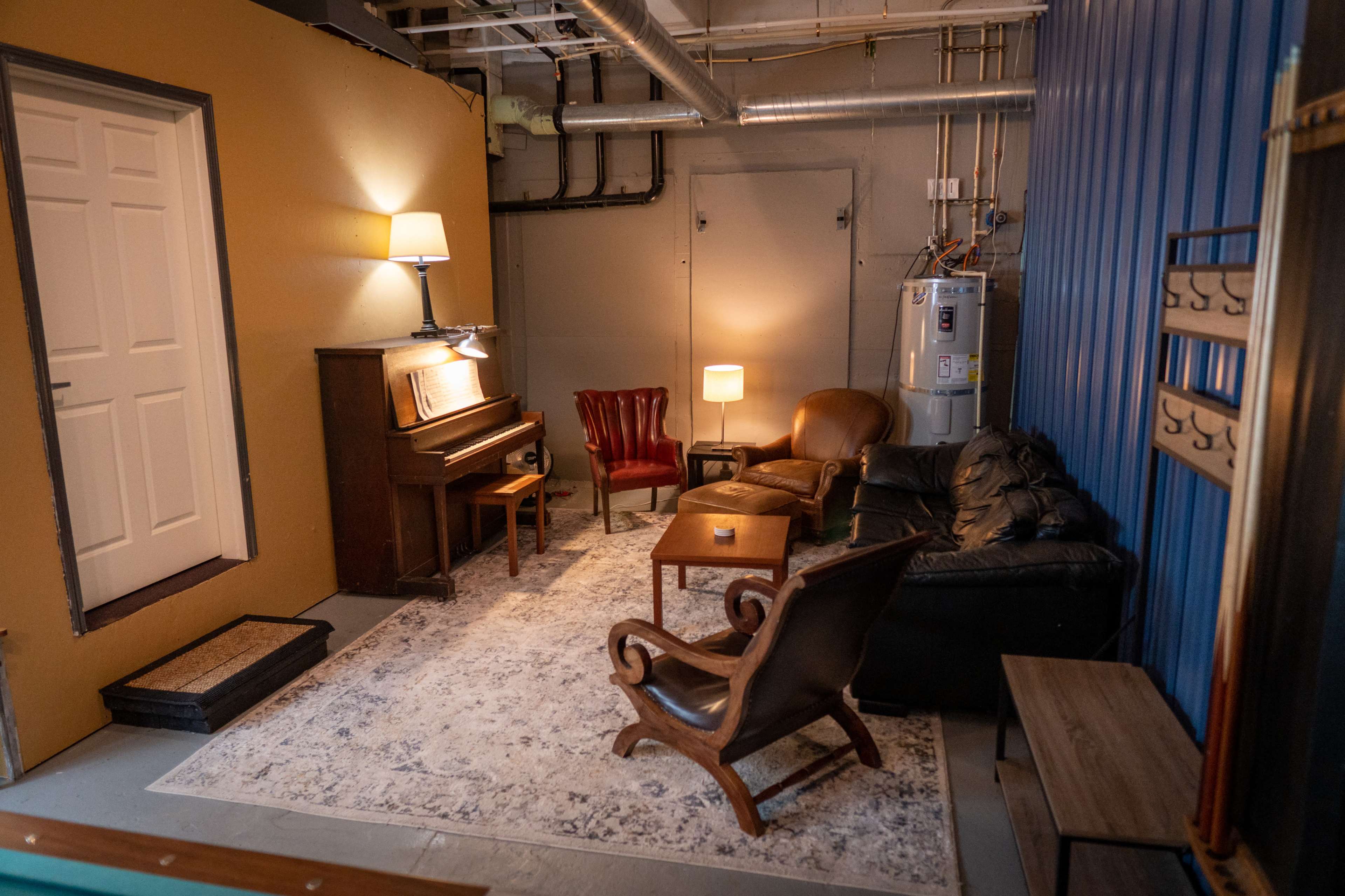 A small, furnished room with a piano, two chairs, a couch, a lamp, and a table on a carpeted floor.
