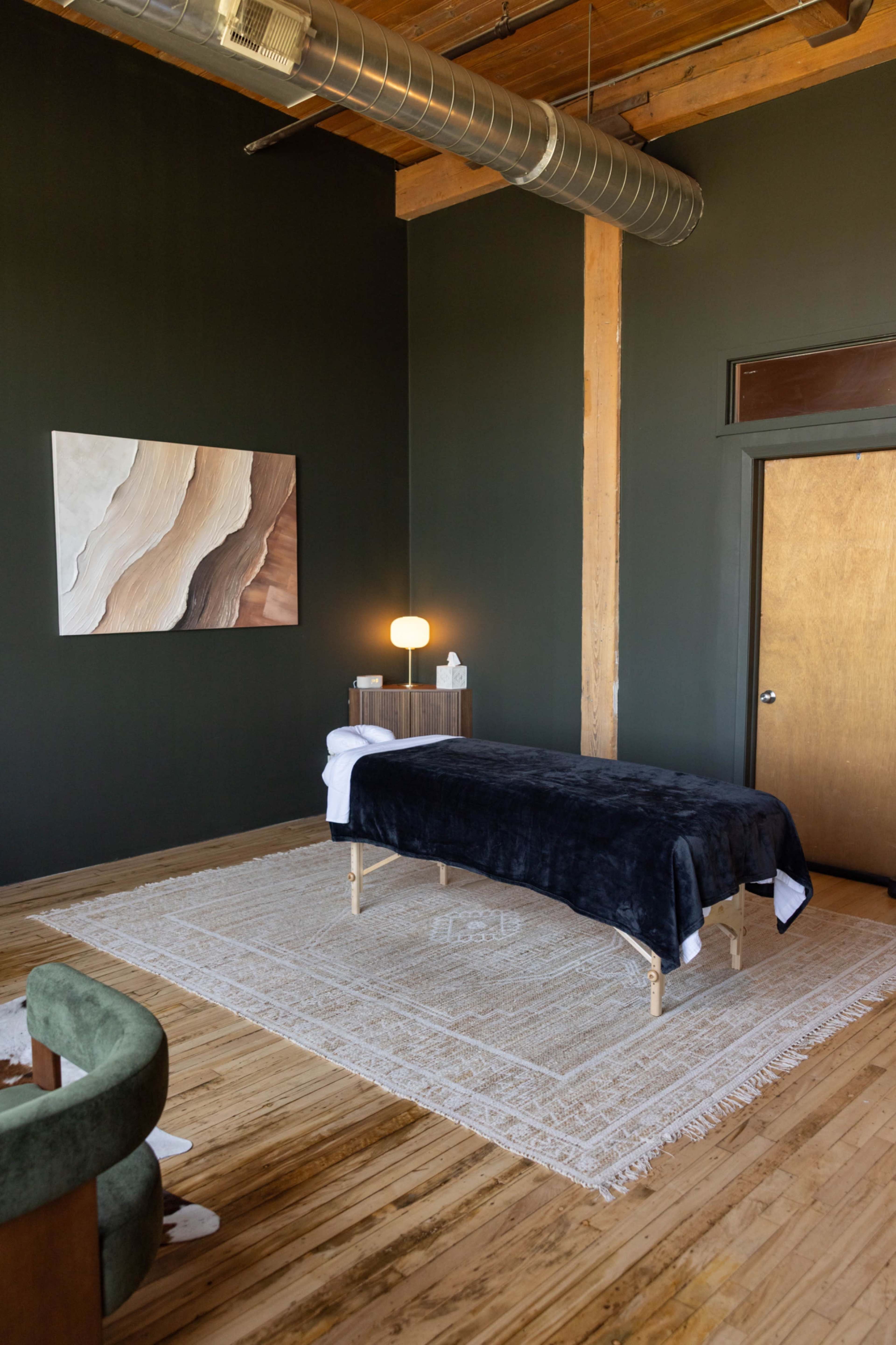 The image shows a serene treatment room with a massage table, minimalist decor, a soft rug, and large windows.