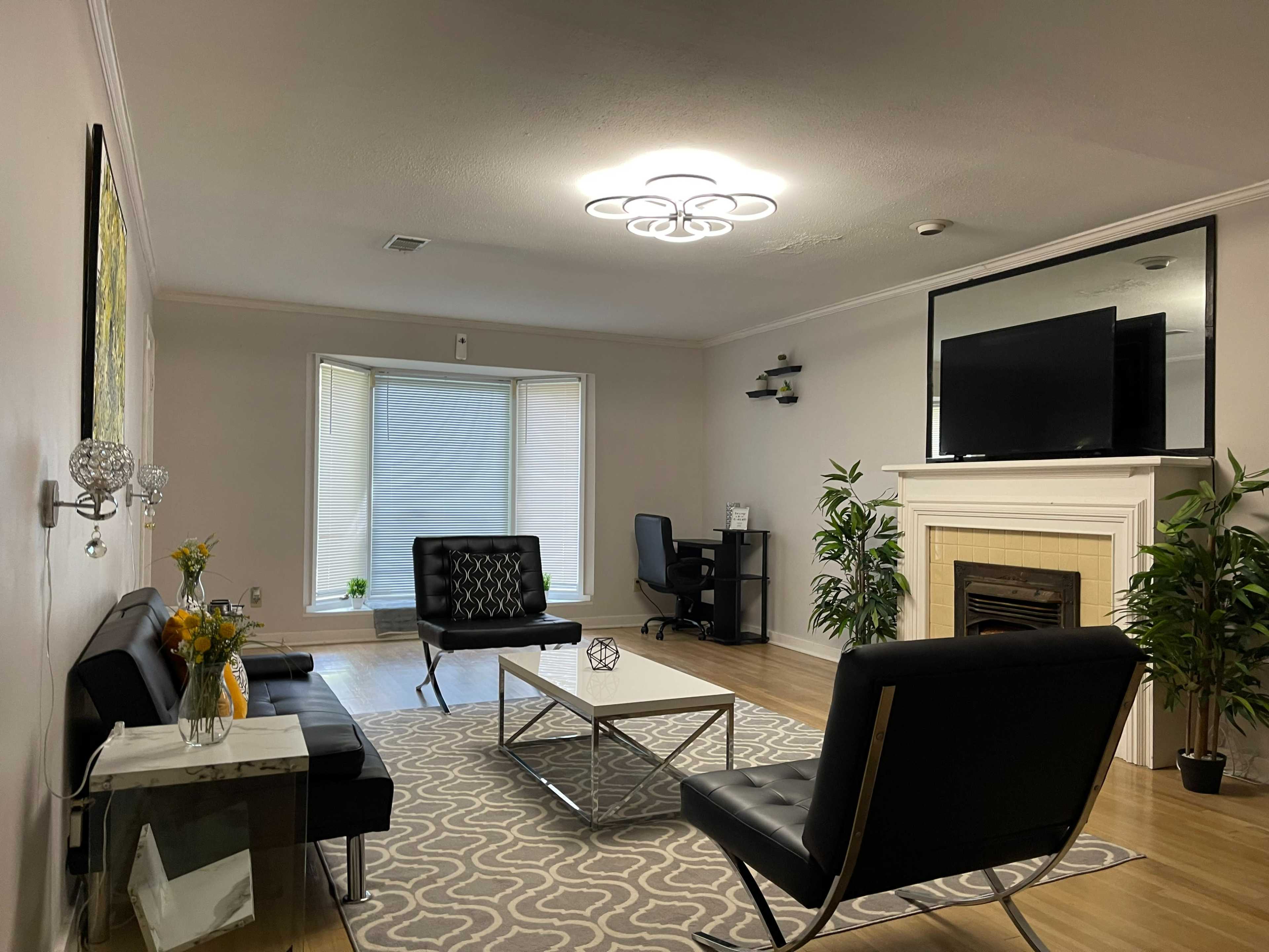 A modern living room featuring a black sofa set, a coffee table on a patterned rug, a fireplace, and a television above it, with a desk in the corner.