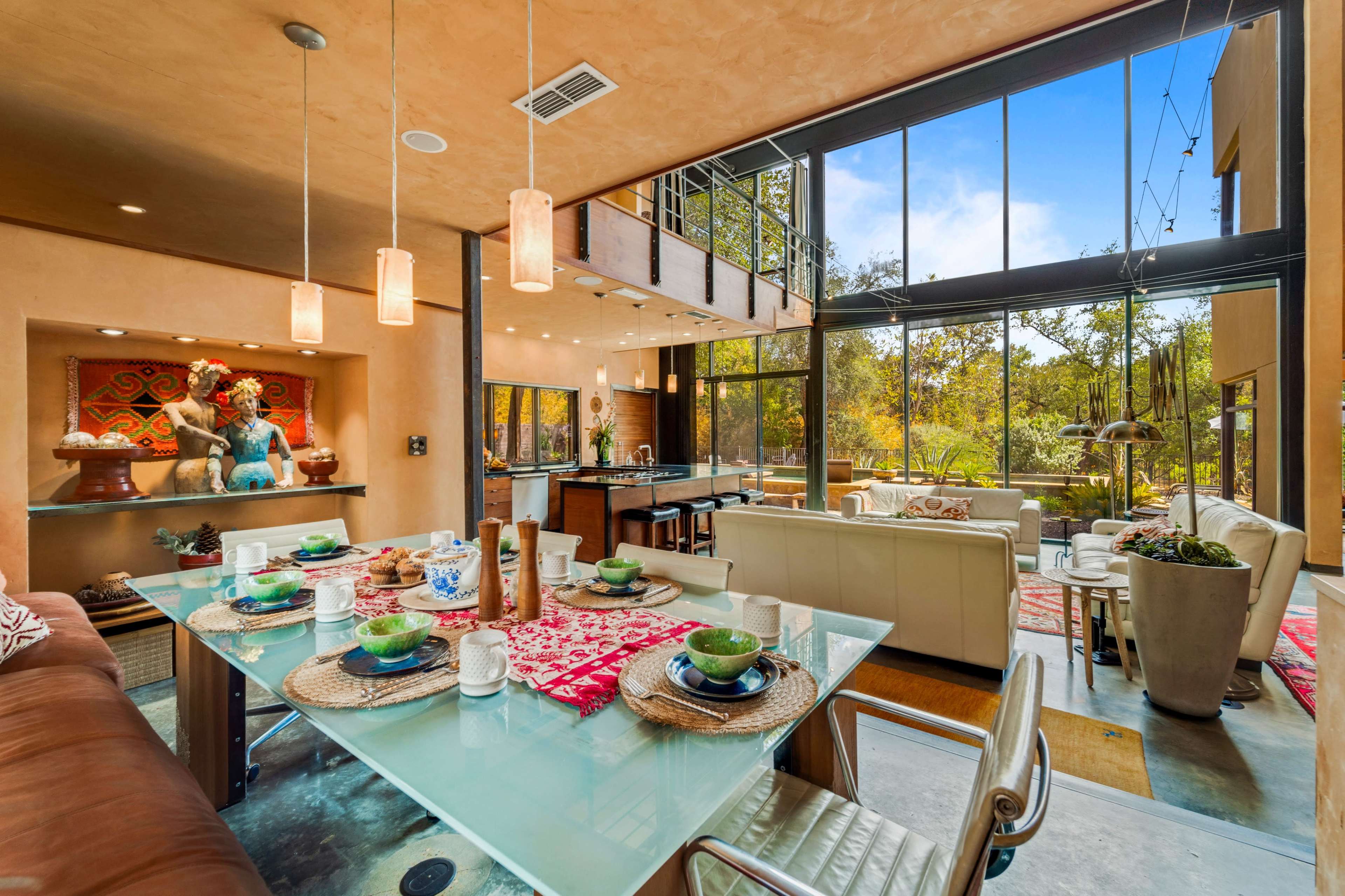 The image shows an open-concept living and dining area featuring large windows that provide a view of the outdoors, with a dining table set for a meal and a modern kitchen in the background.