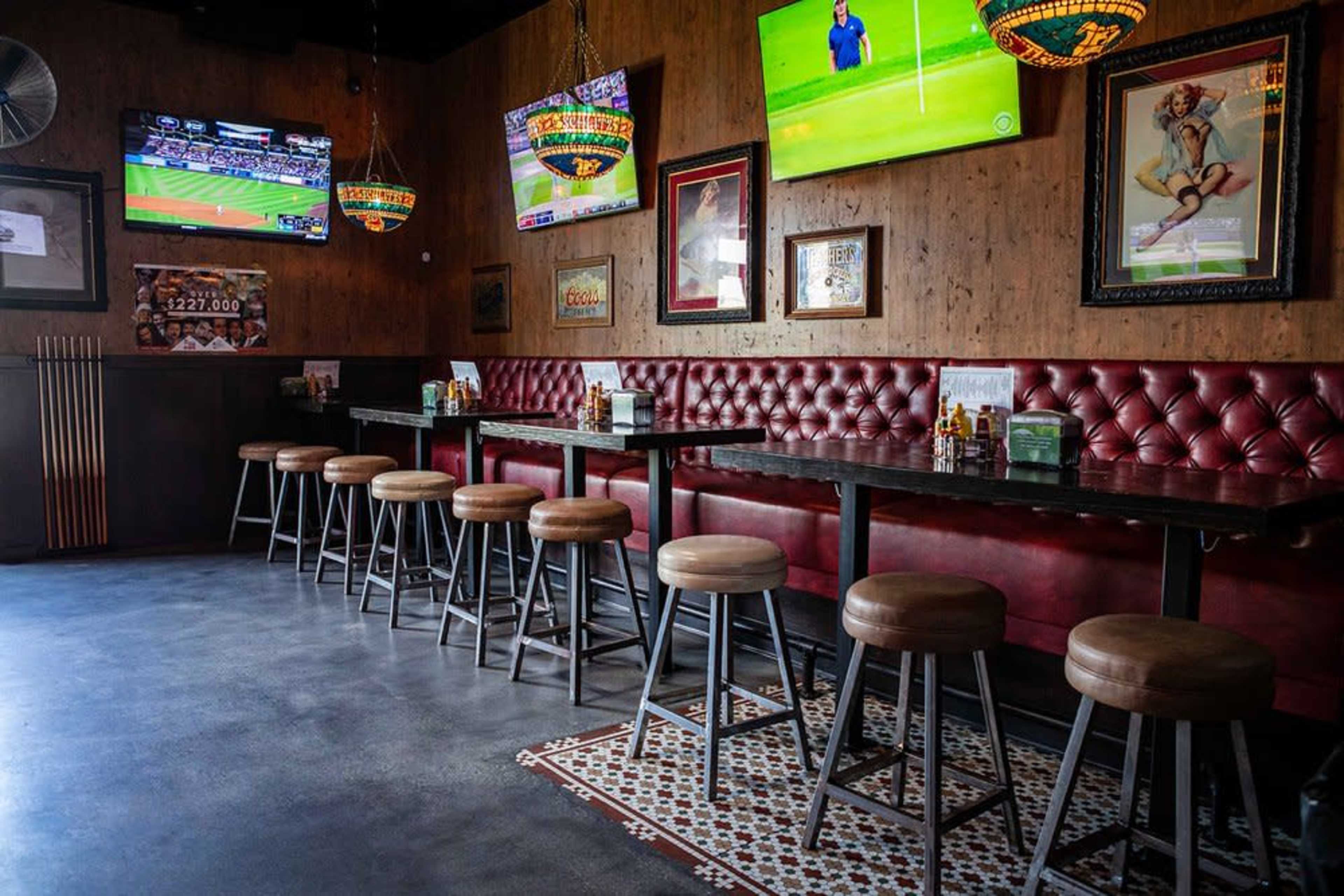 The image shows an empty bar area with several TV screens mounted on the walls, red upholstered seating along the back, and wooden stools arranged in front of dark tables.