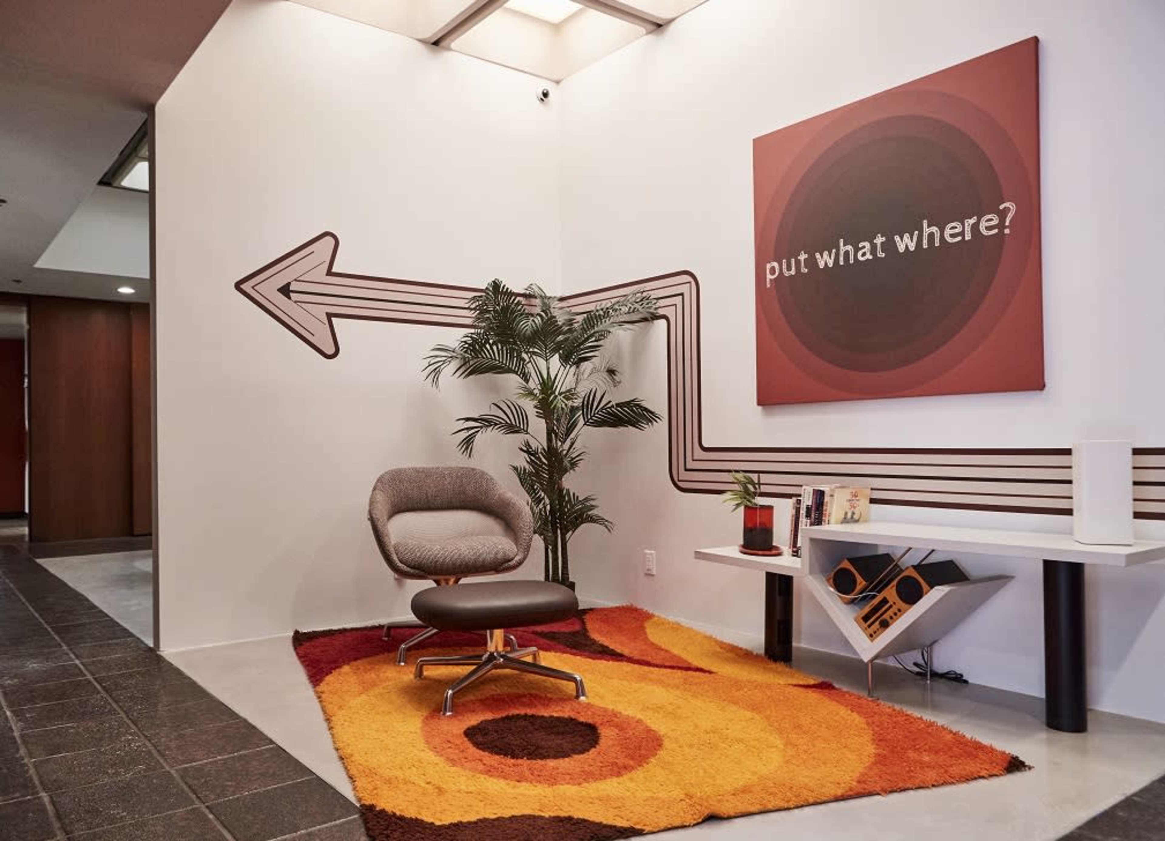 A modern entryway with a chair, a colorful area rug, a potted plant, and a wall art piece that includes an arrow and the phrase "put what where?"
