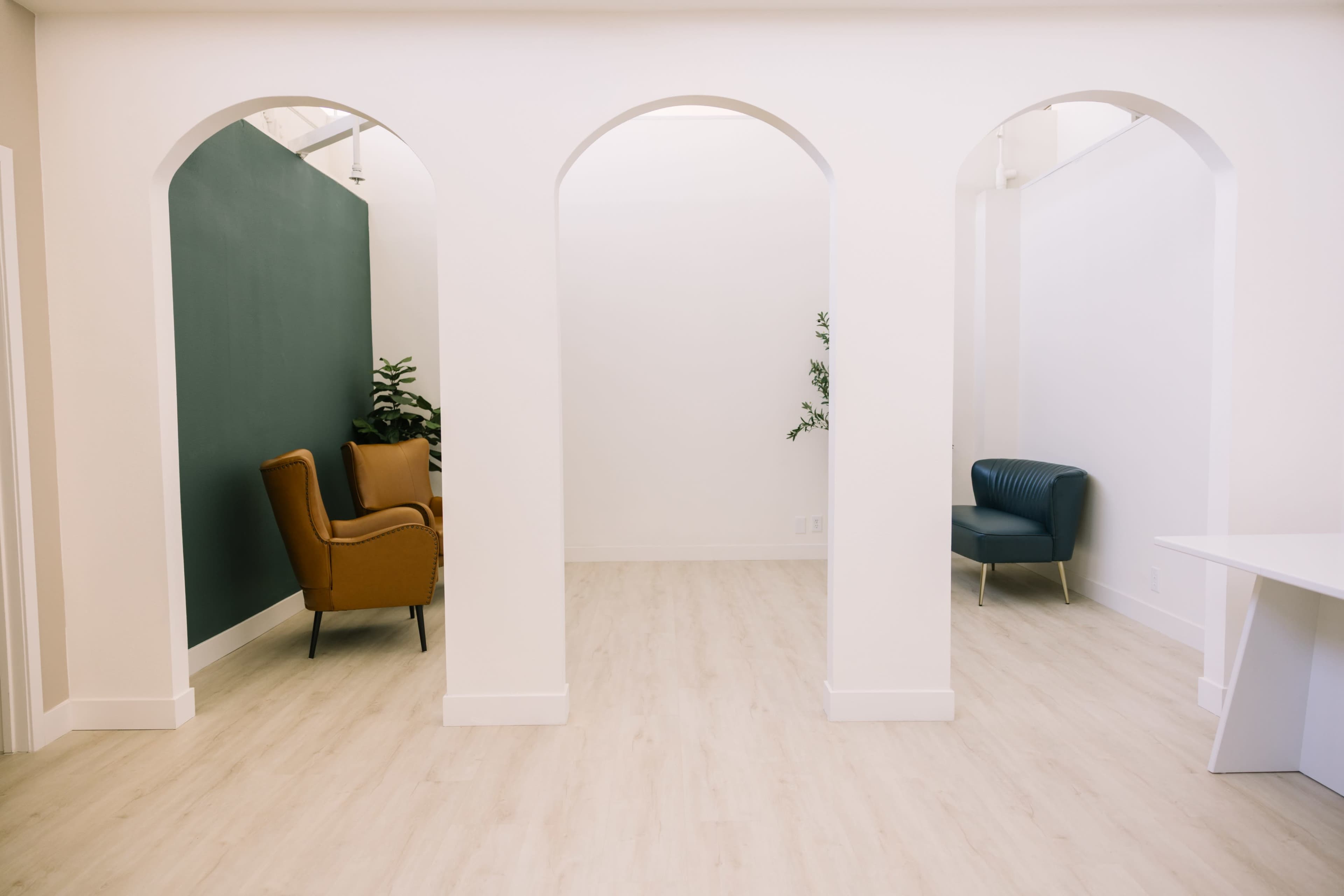 The image shows a minimalist interior with three arched openings, a light-colored wooden floor, and two accent chairs placed in a corner.