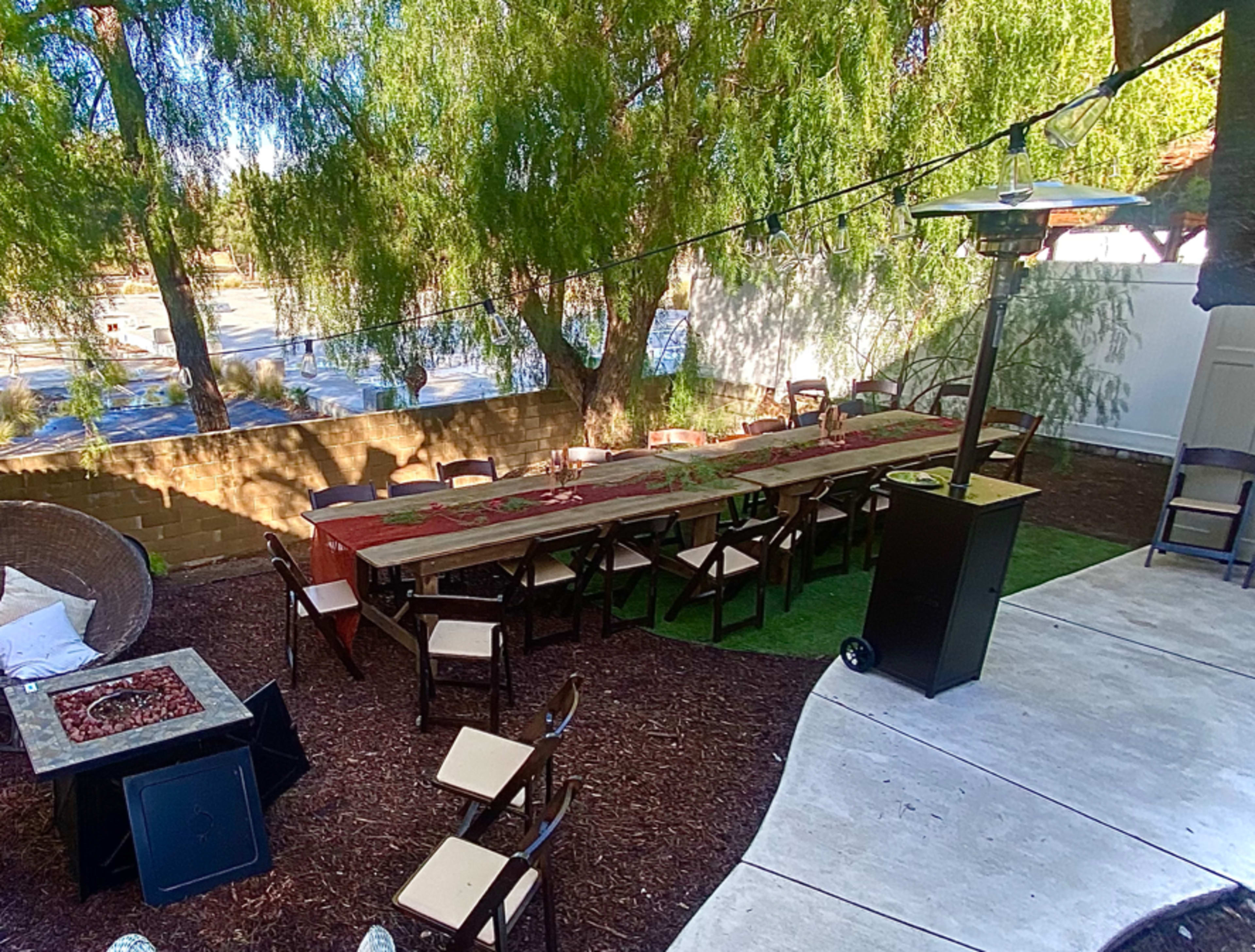A long wooden table with chairs is set up outside under a large tree, accompanied by a patio heater and a seating area made of woven material.