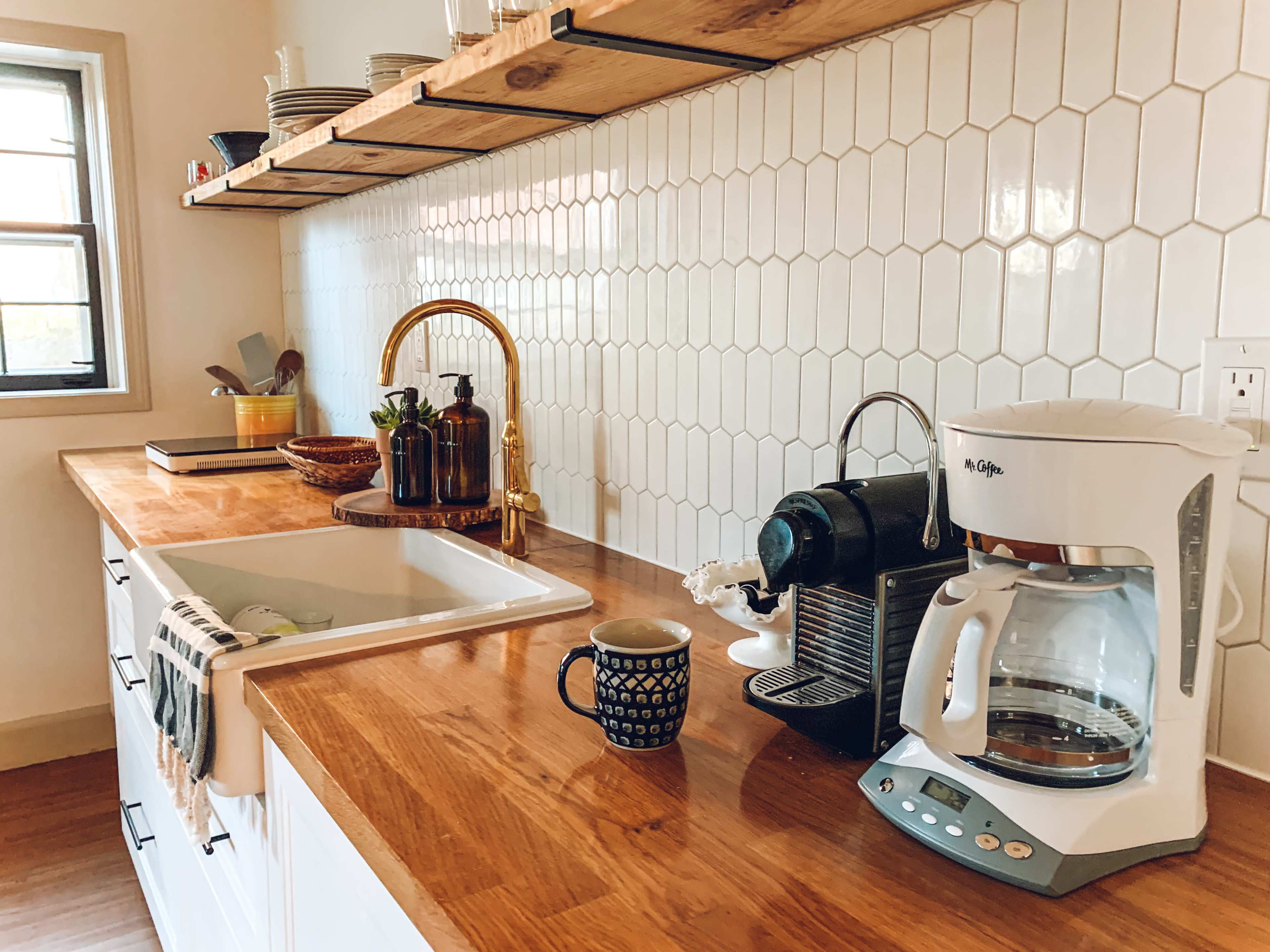 A kitchen countertop features a coffee maker, a kettle, and a mug next to a sink, with a backsplash of hexagonal white tiles and wooden shelves above displaying dishes.