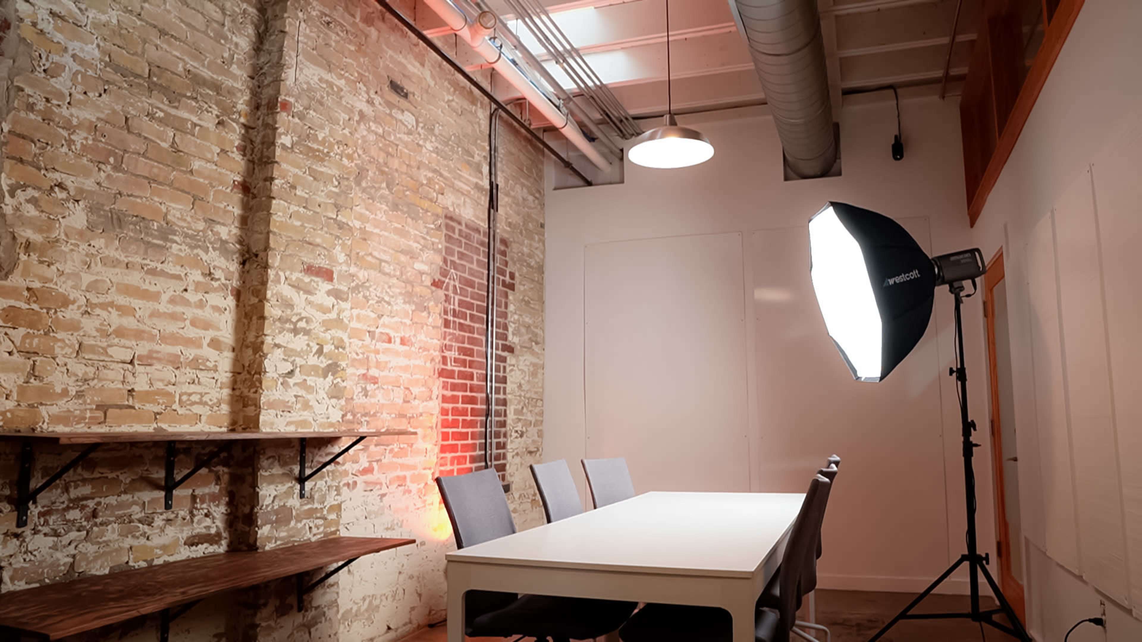 A spacious meeting room features a white table surrounded by gray chairs, with exposed brick walls and professional lighting equipment set up in the corner.