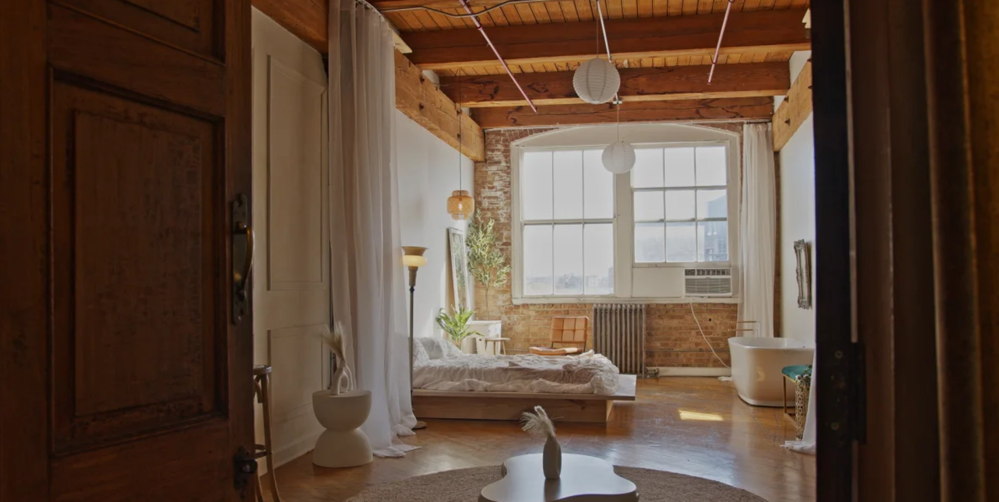 The image shows a spacious, well-lit bedroom with exposed wooden beams, a large window, and a mix of modern and rustic decor.