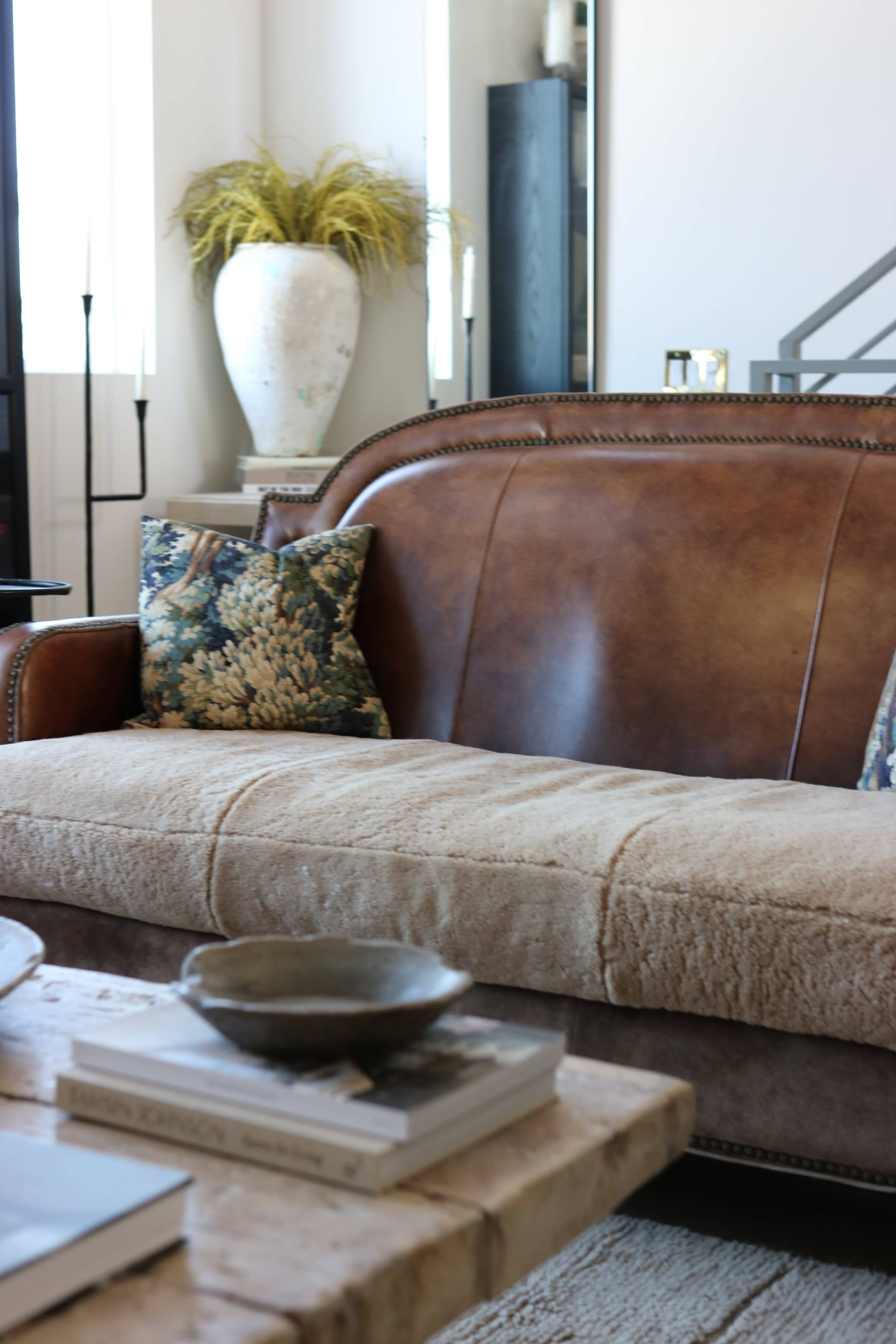 The image features a leather sofa with decorative pillows, accompanied by a wooden coffee table and a large ceramic vase in a well-lit living room.