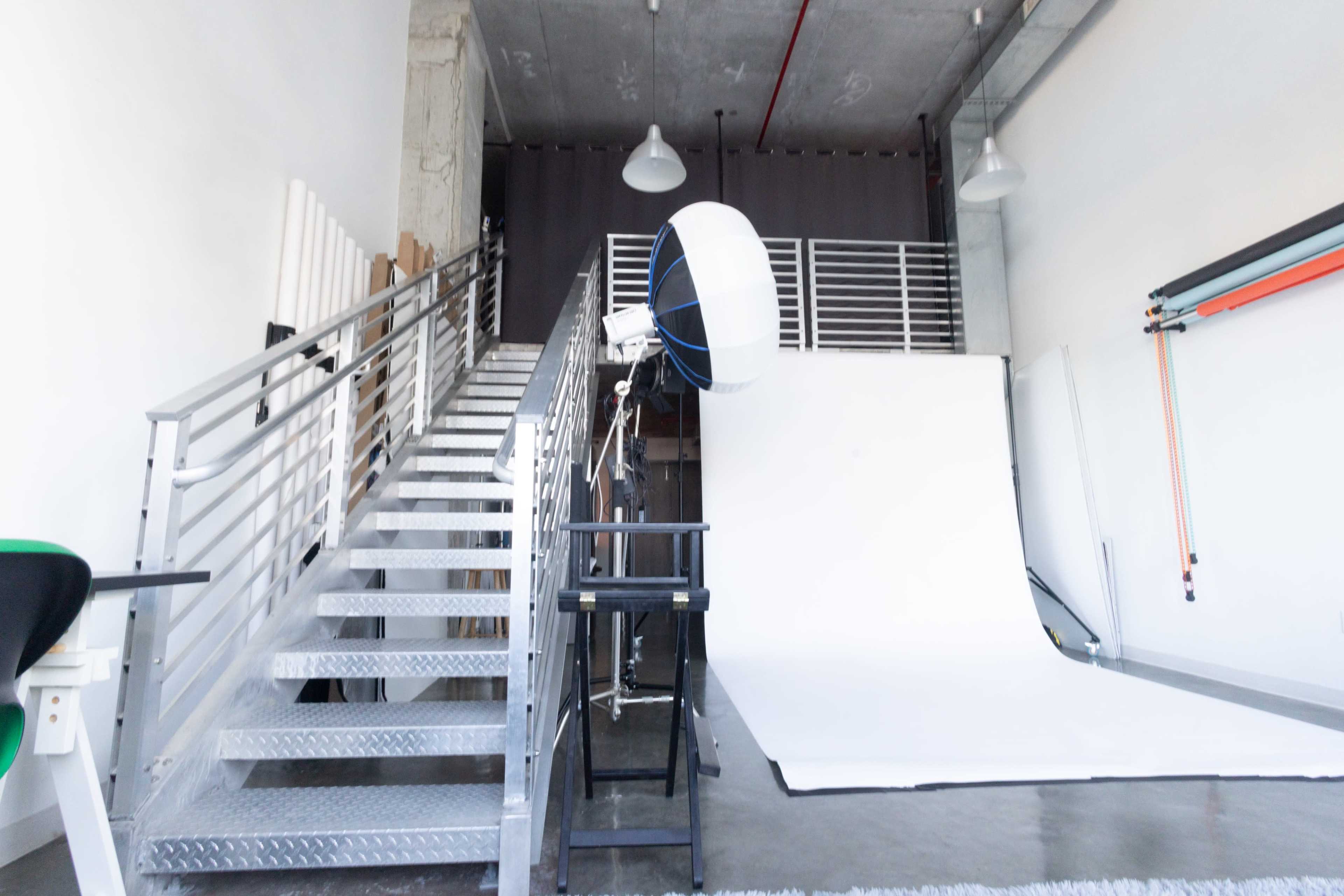 The image shows a modern photography studio featuring a staircase leading to an elevated area, with a white backdrop and lighting equipment set up.