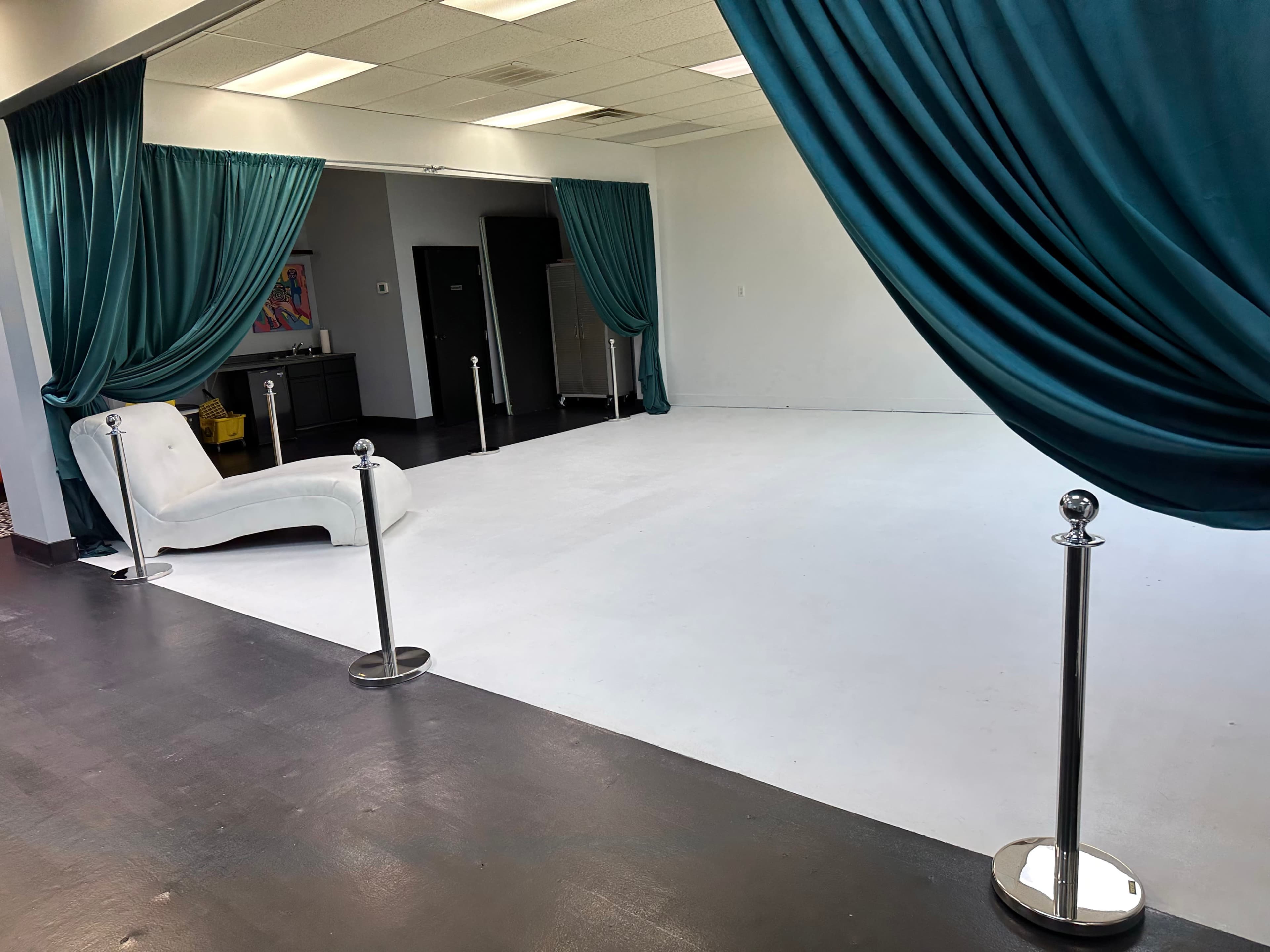 A spacious studio with white walls and floor, featuring a white curved sofa, teal curtains, and stanchions with ropes.