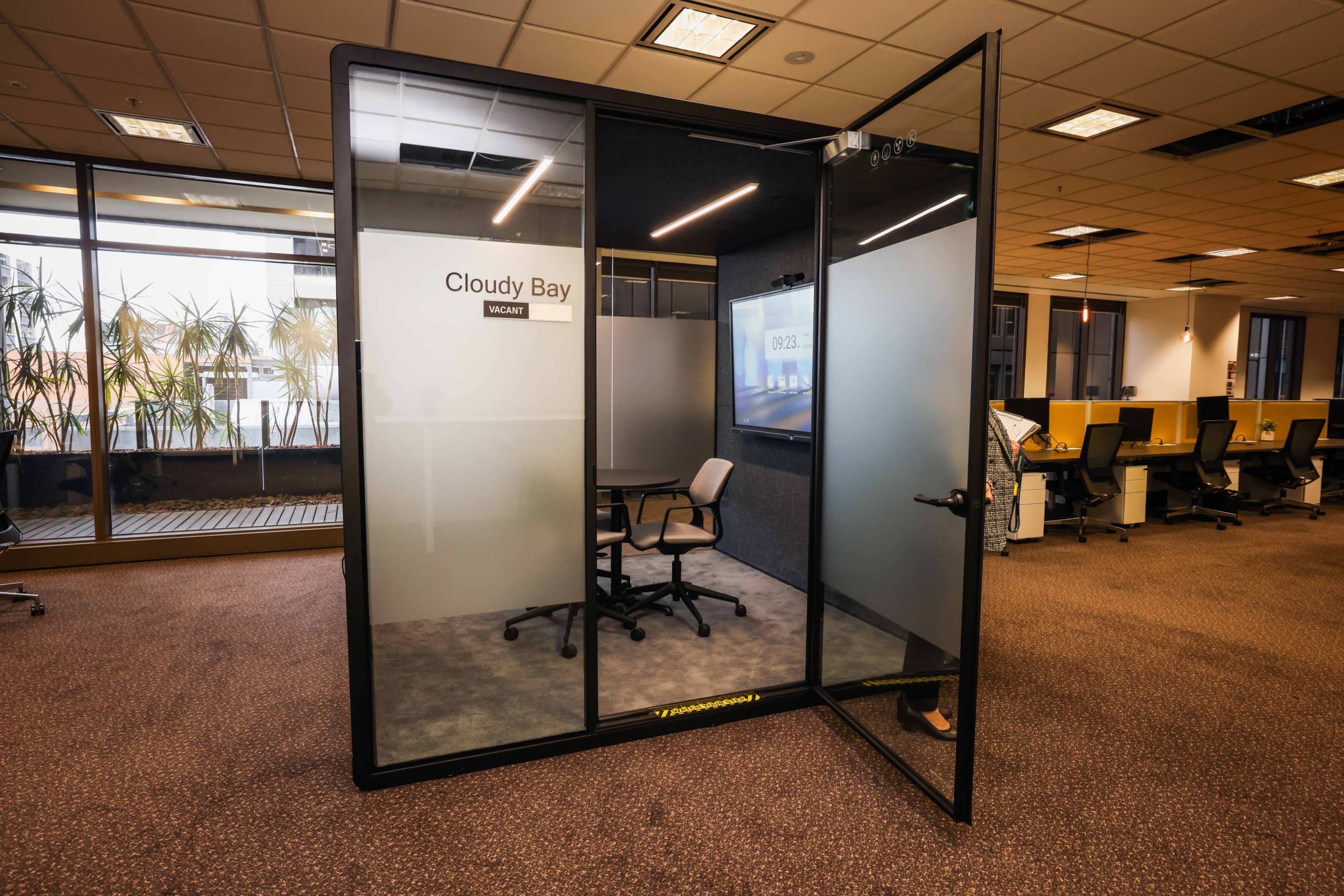 The image shows a glass-walled conference room labeled "Cloudy Bay," equipped with a single chair and a presentation screen, situated in an open office space with plants in the background.