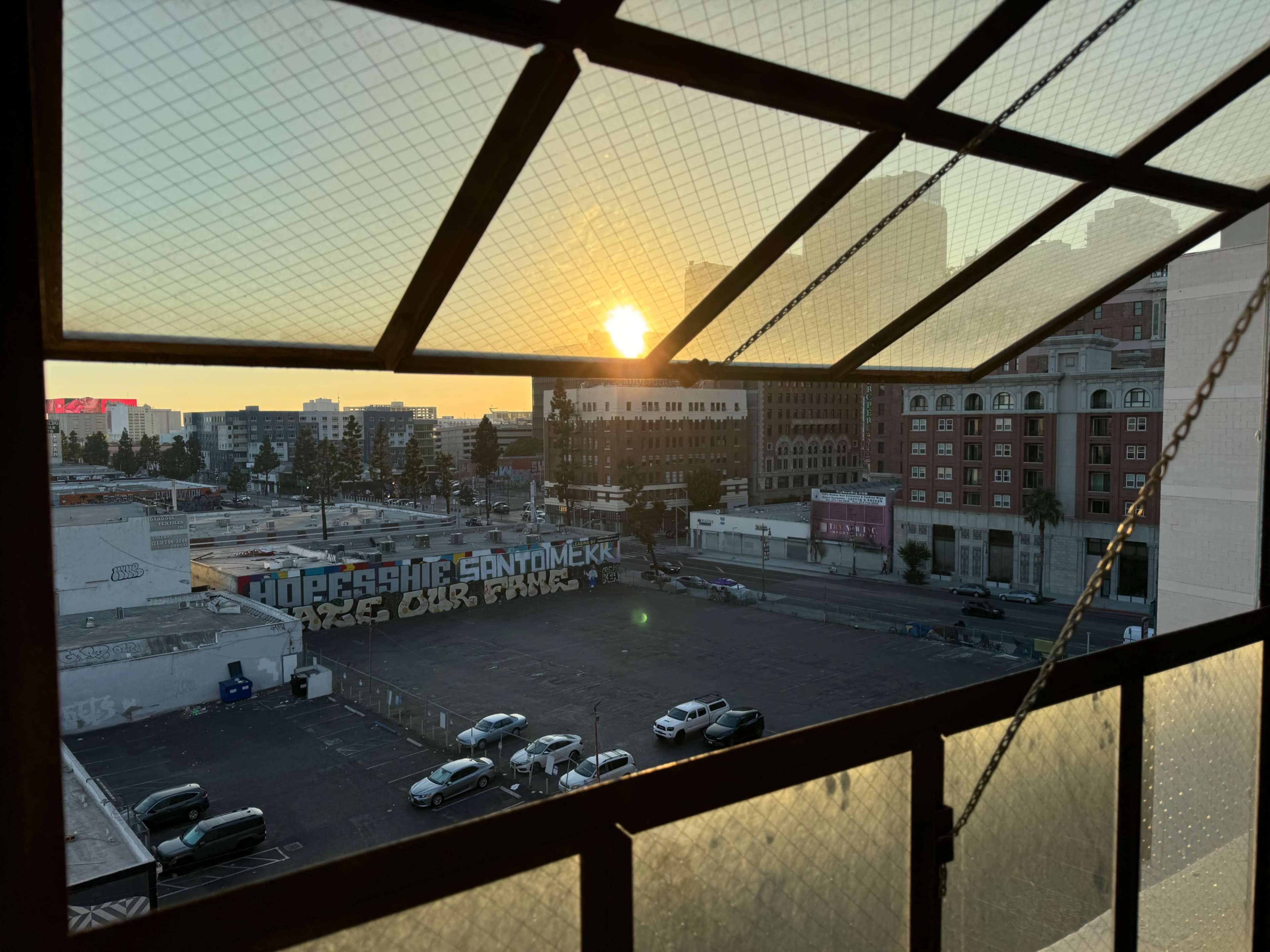 The sun rises over a cityscape, illuminating parked cars and buildings visible through a window's grid.