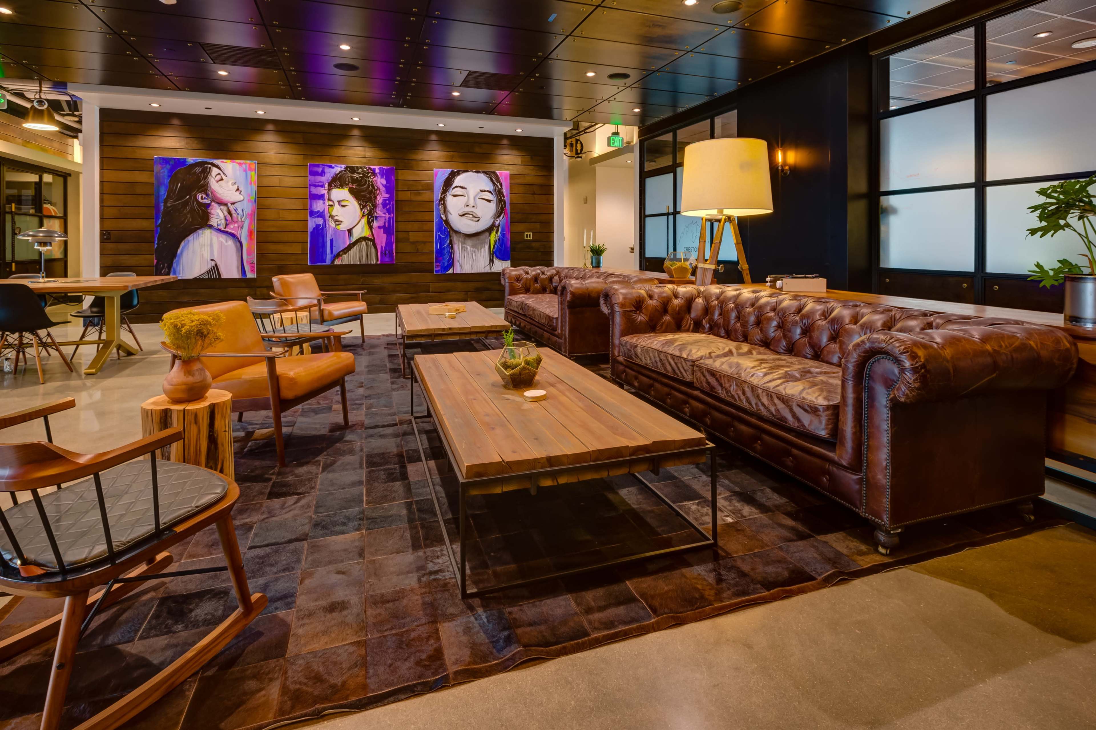 A modern lounge area featuring a brown leather chesterfield sofa, wooden tables, art on the walls, and a mix of seating options.