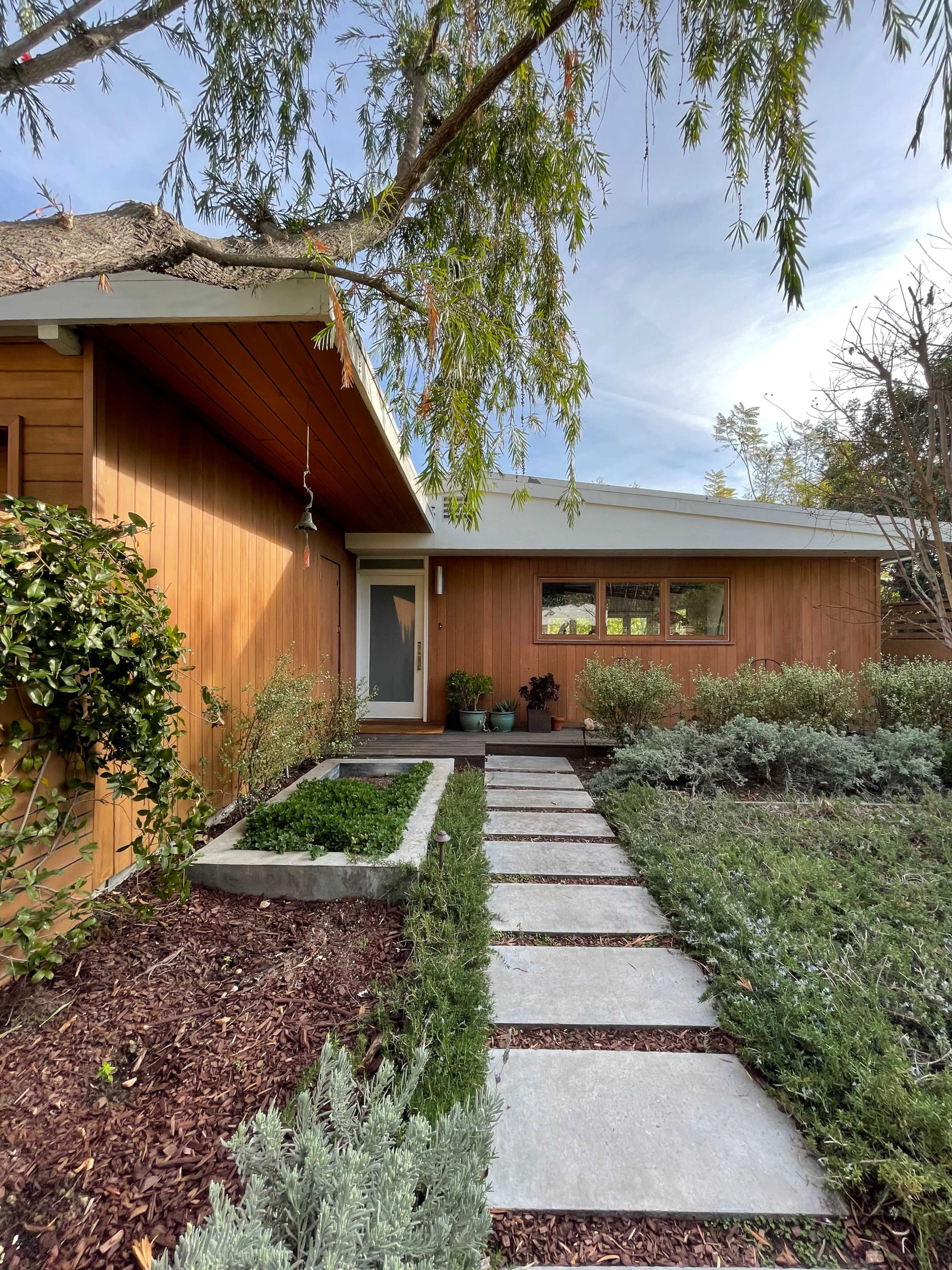 A modern wooden house features a landscaped pathway of concrete slabs surrounded by greenery and mulch.