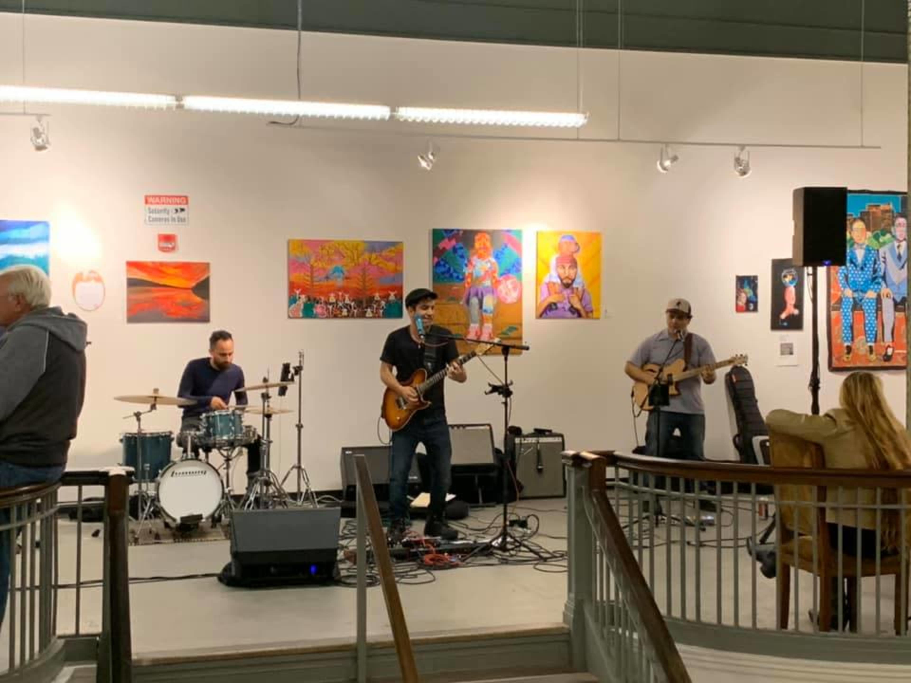 A band performs on a small stage in a gallery surrounded by colorful artwork and an audience observing the performance.