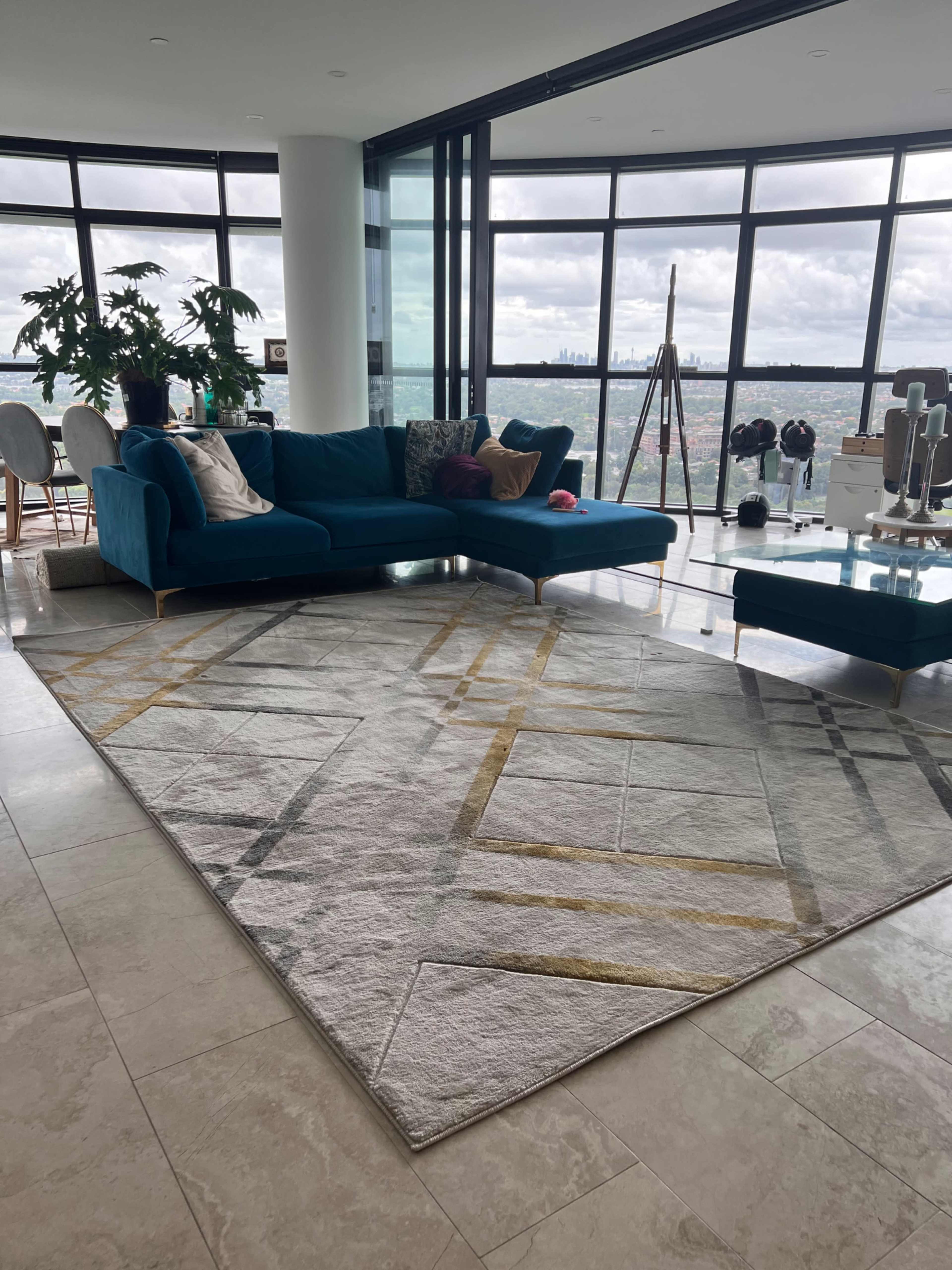 The image shows a contemporary living room with a large turquoise sectional sofa, a geometric area rug, and large windows offering a city view.