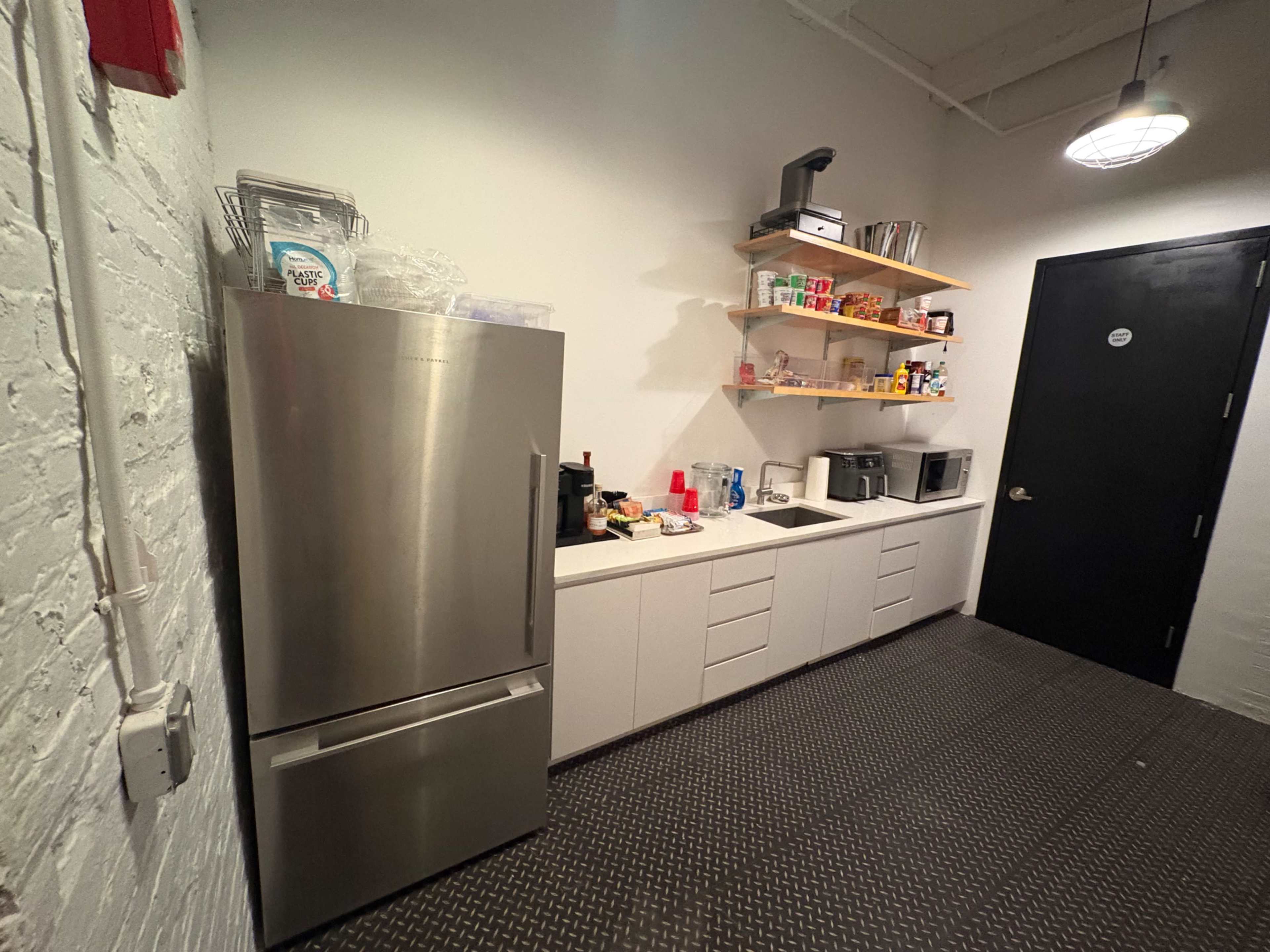 The image shows a modern kitchen with a stainless steel refrigerator, open shelves filled with various food items and kitchen supplies, and a dark door at the end of the space.
