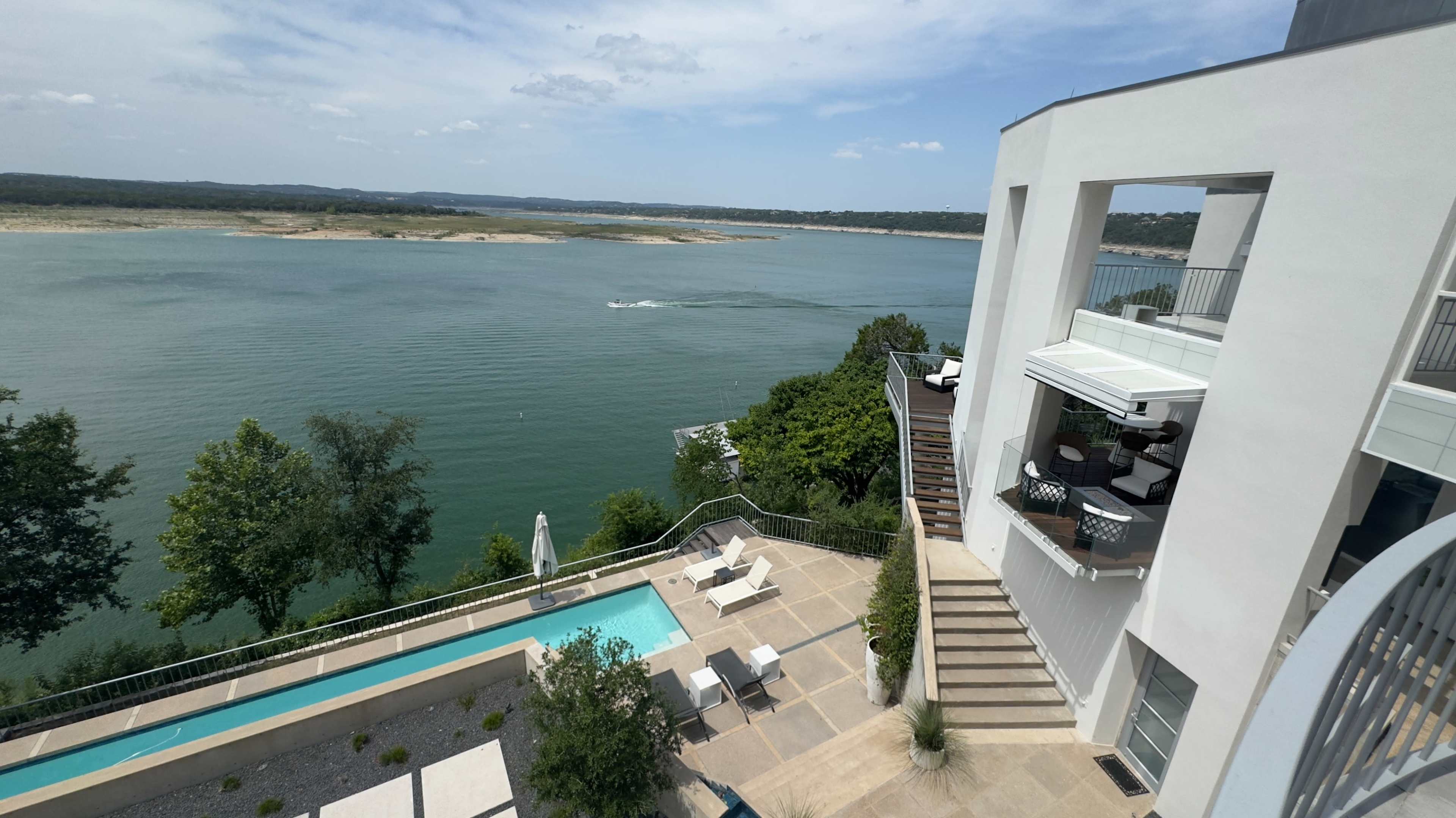 A modern white building with a swimming pool overlooks a serene lake, surrounded by greenery and hills in the distance.