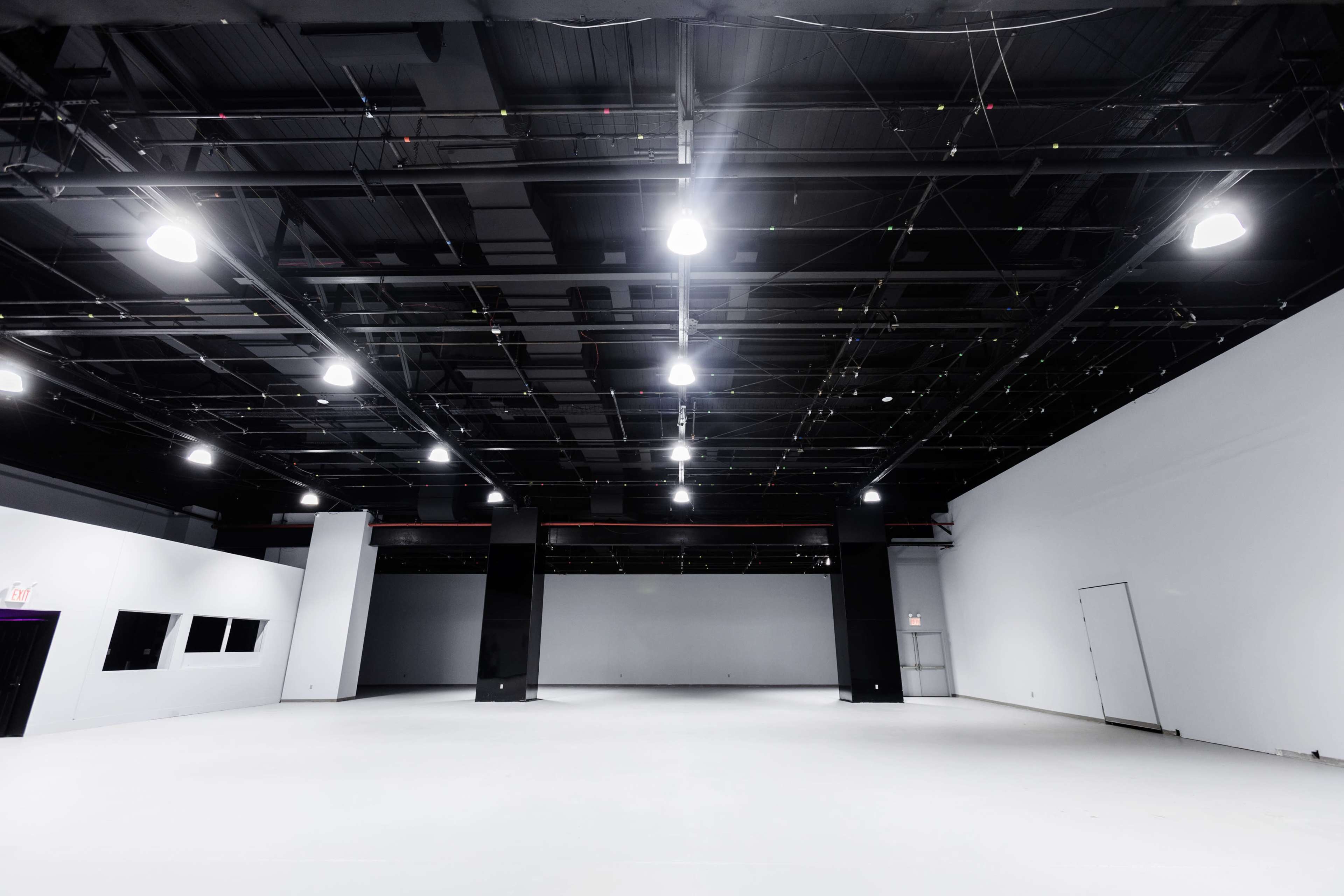 The image shows a large, empty warehouse space with white walls and a ceiling filled with hanging lights and cables.