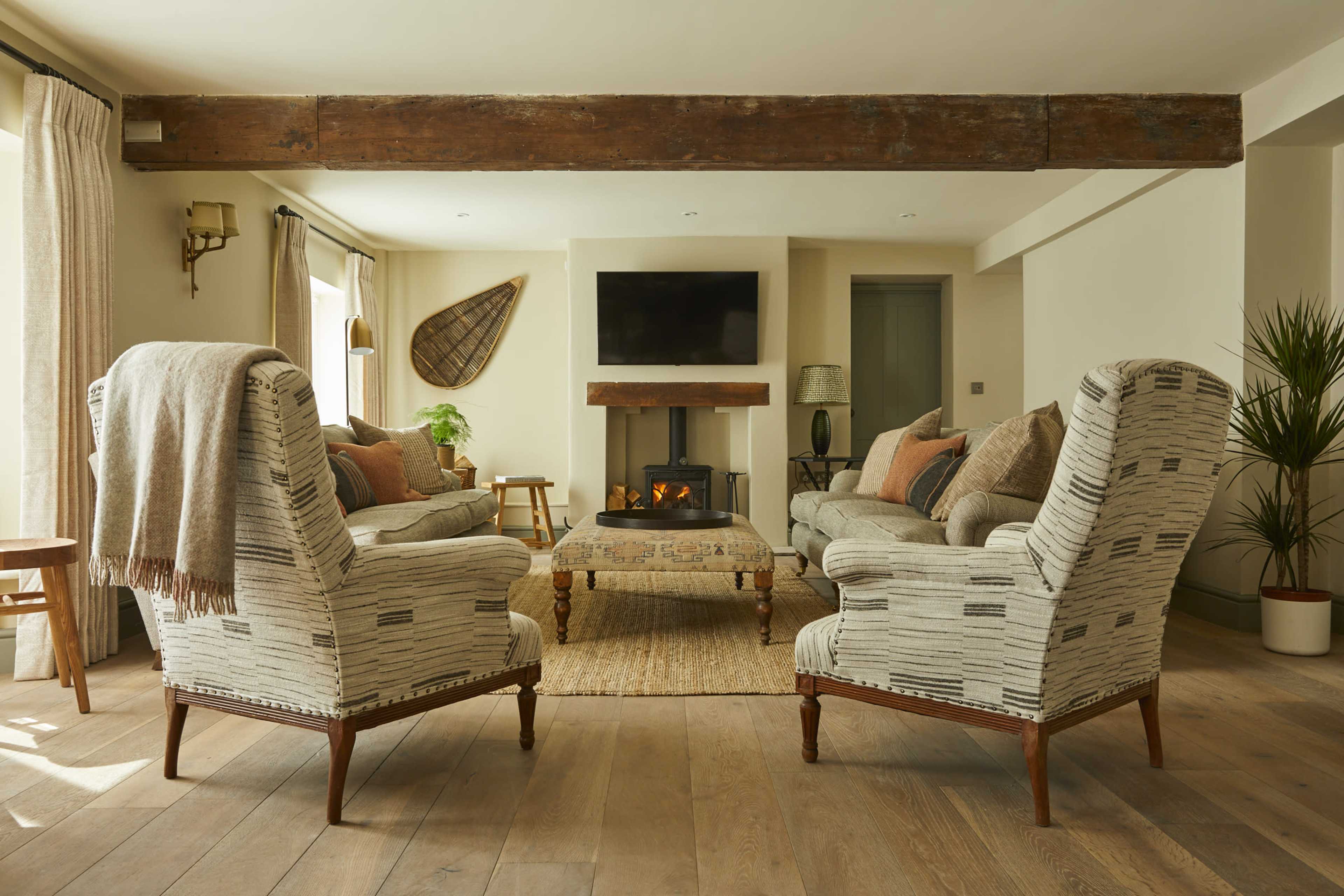 A spacious living room features two patterned armchairs and a sofa, arranged around a central coffee table in front of a fireplace, with natural light streaming through large windows.