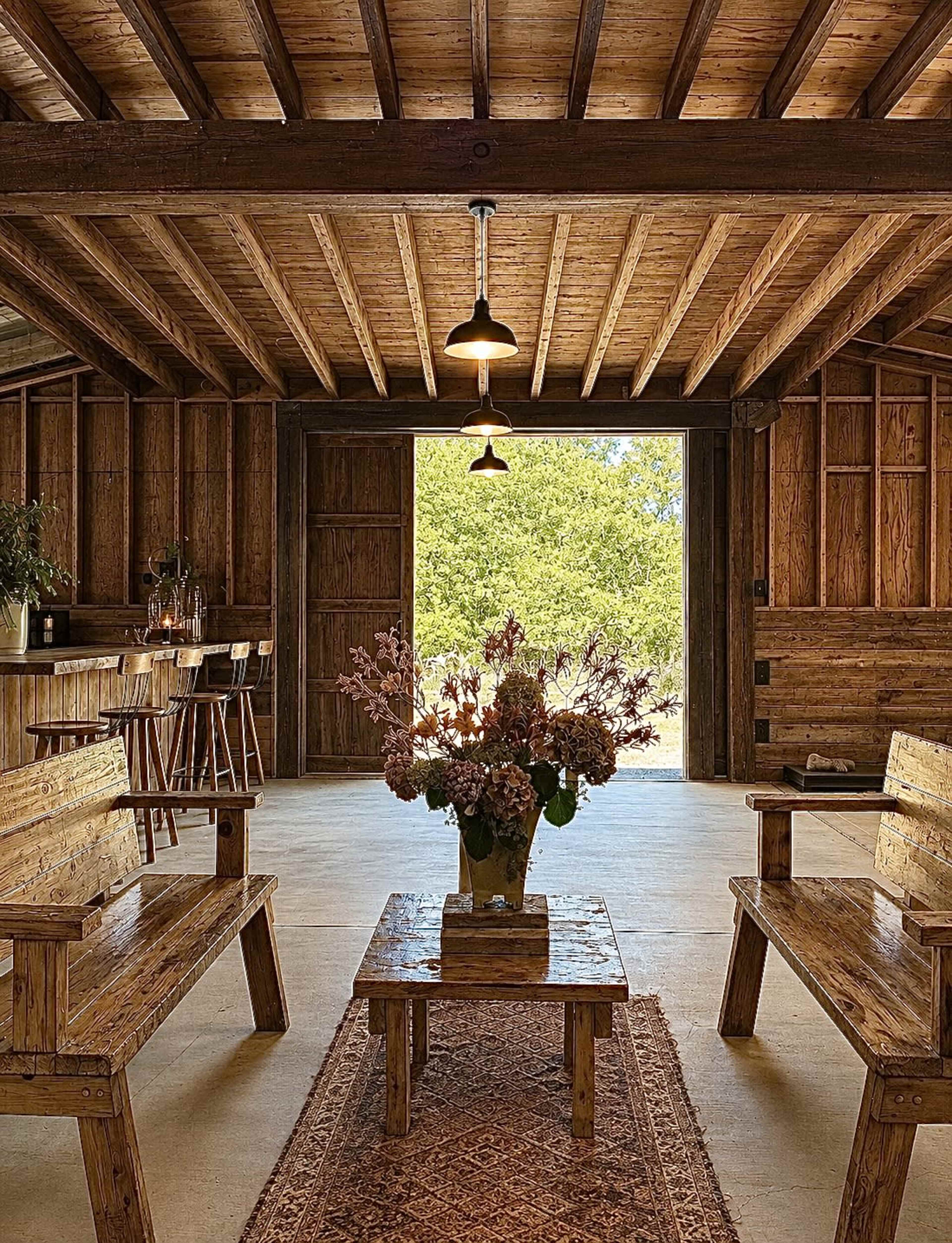 The image features a rustic interior of a wooden barn, showcasing wooden benches and a central table adorned with flowers, while large doors open to a view of greenery outside.