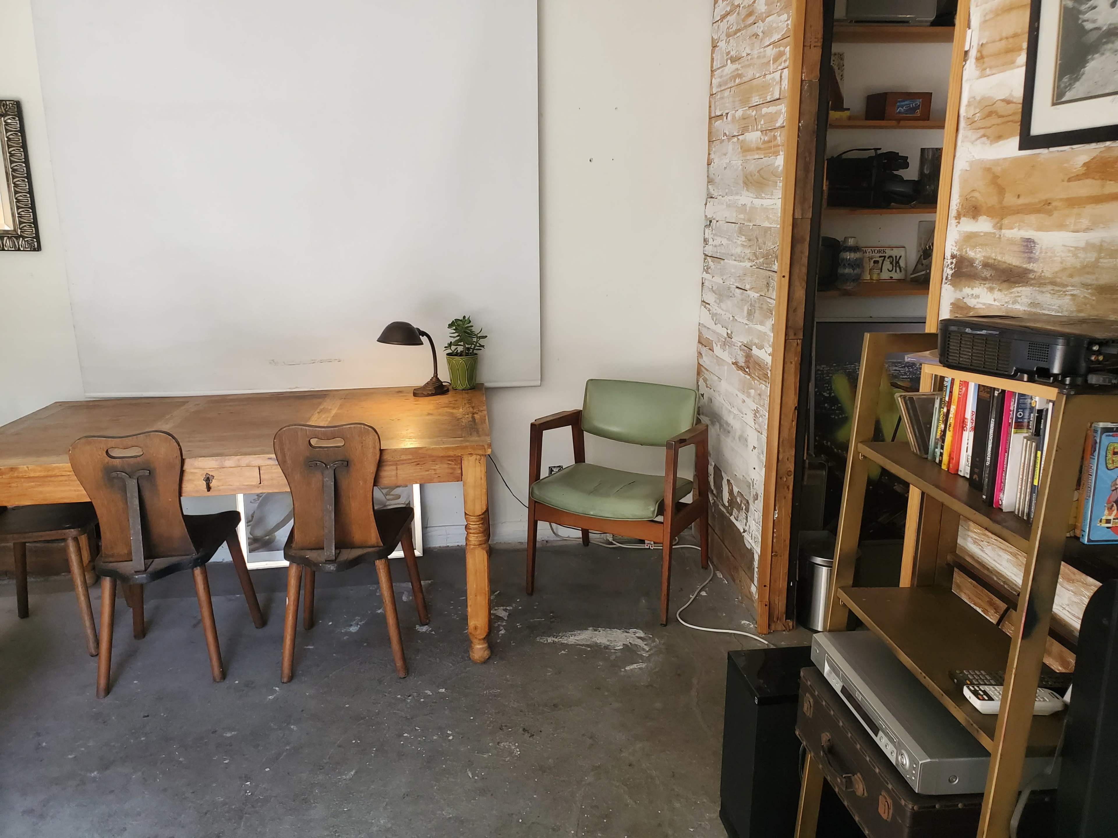 The image shows a rustic workspace featuring a wooden table with three chairs, a small lamp, and a green armchair, along with a bookshelf filled with books and a projector in the corner.
