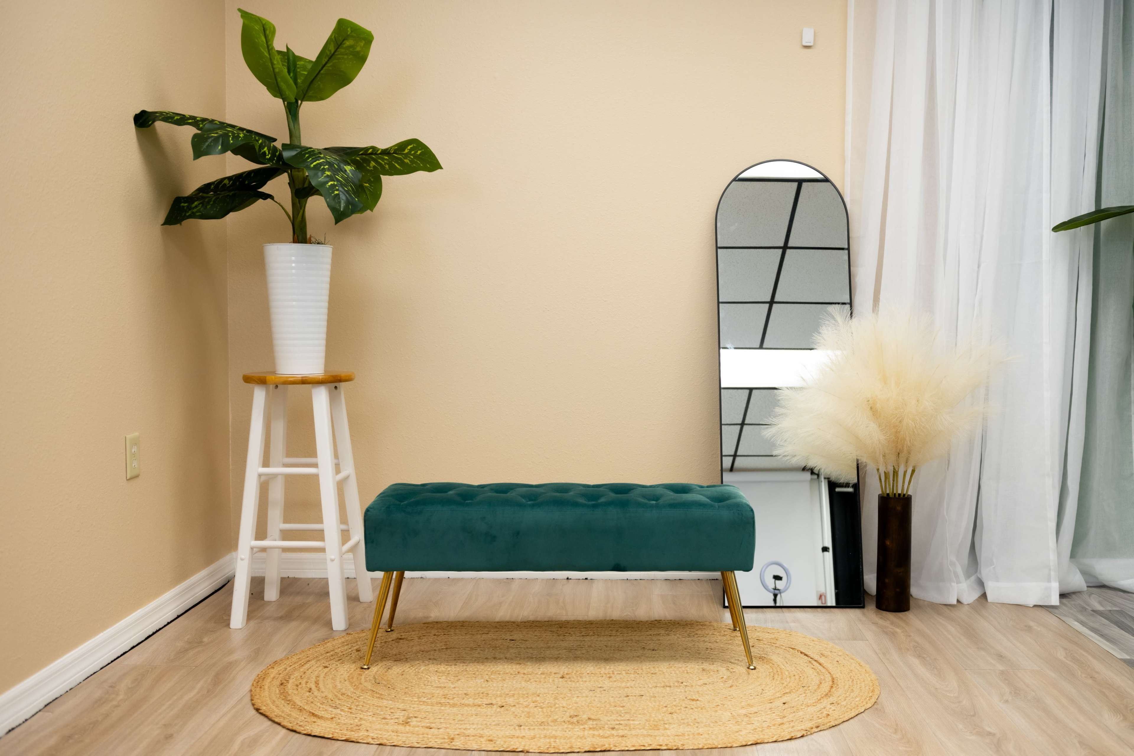 The image features a light-colored room with a green upholstered bench, a tall plant in a white pot, a round woven rug, and a mirror reflecting part of the space.