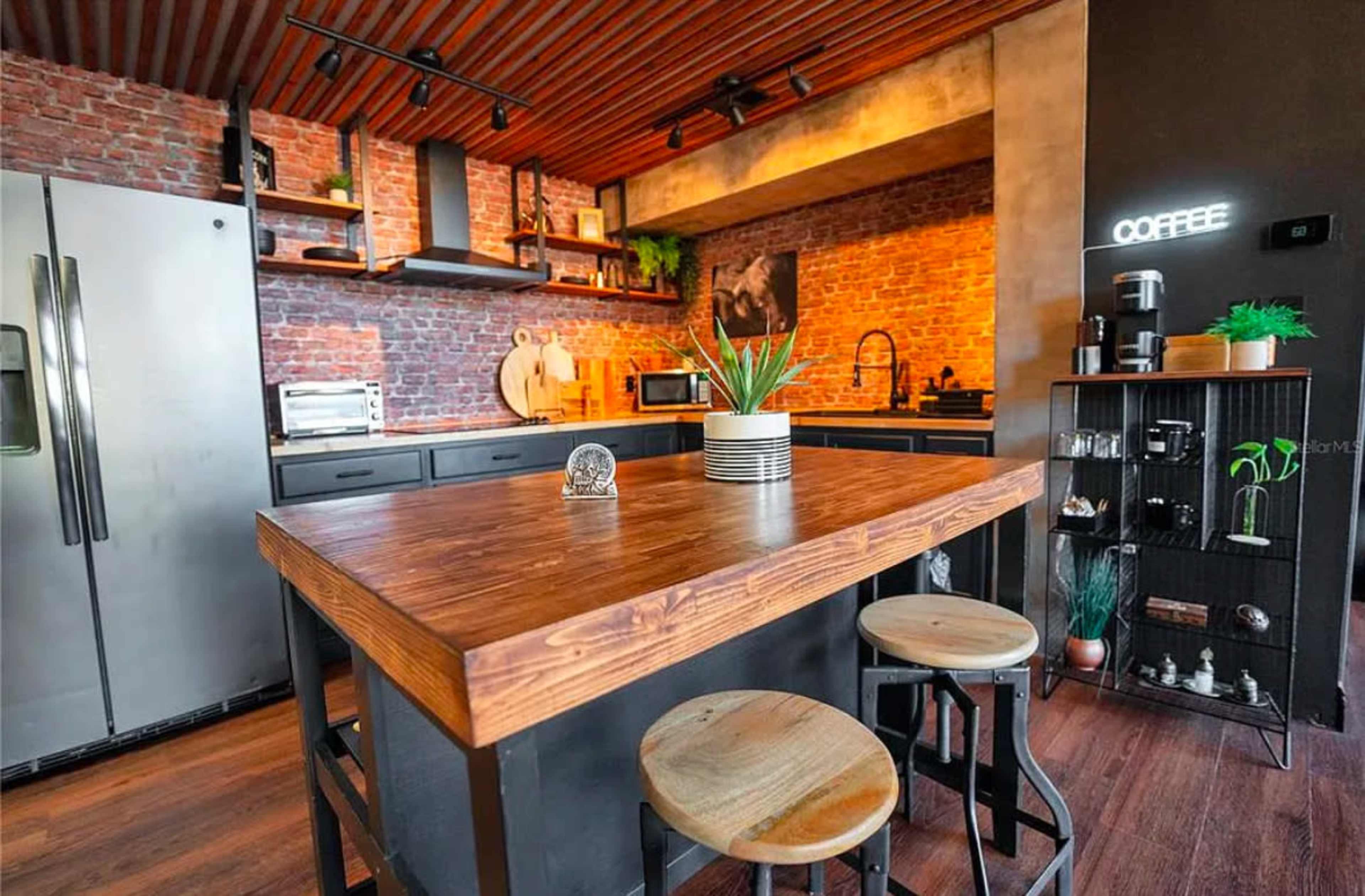 The image shows a modern kitchen featuring a central wooden island with stools, exposed brick walls, and stainless steel appliances.