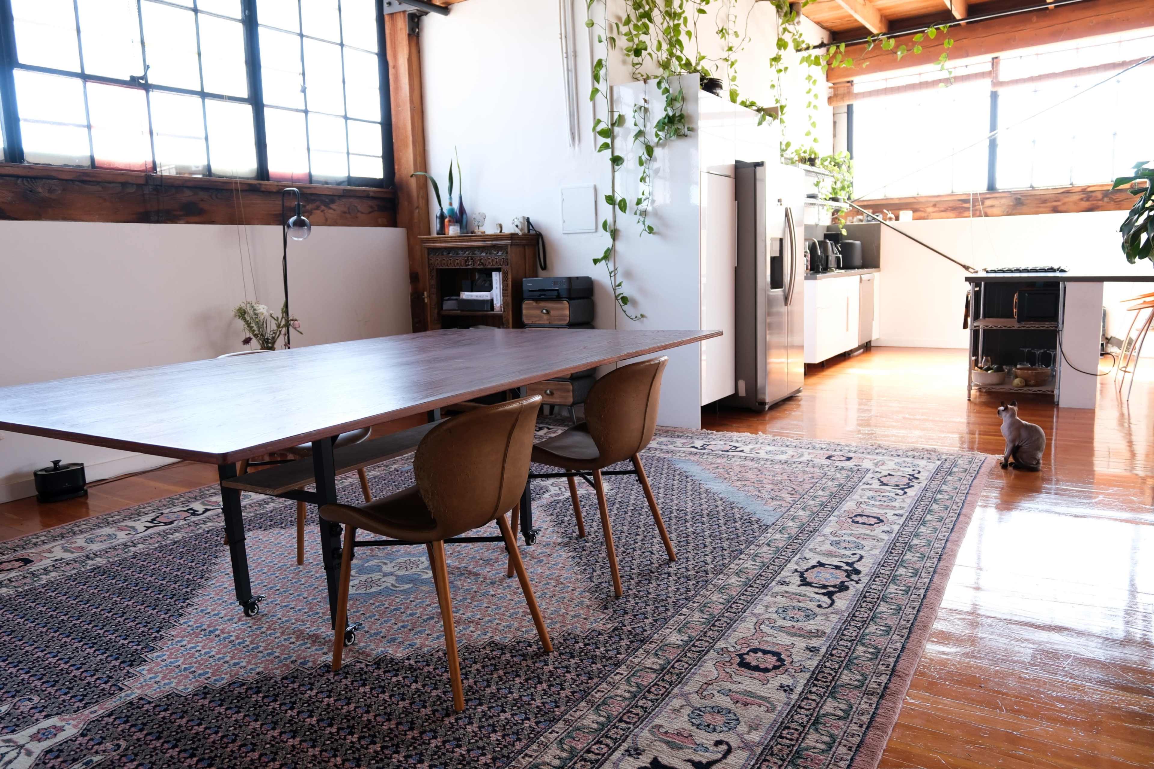 A spacious dining area features a large wooden table with two chairs, surrounded by a patterned rug and plants, adjacent to a modern kitchen with a refrigerator and appliances.