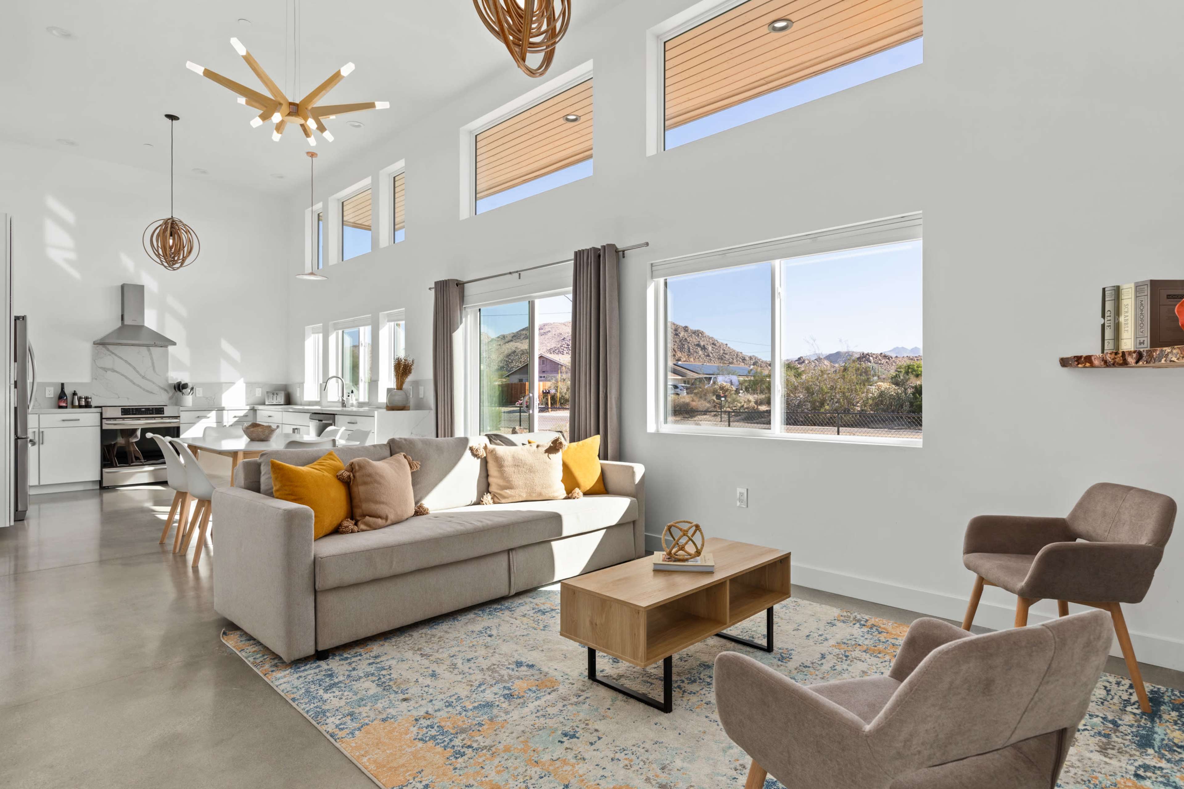 A modern living room features a light-colored sofa with yellow accent pillows, a wooden coffee table, and large windows providing a view of the outdoors.