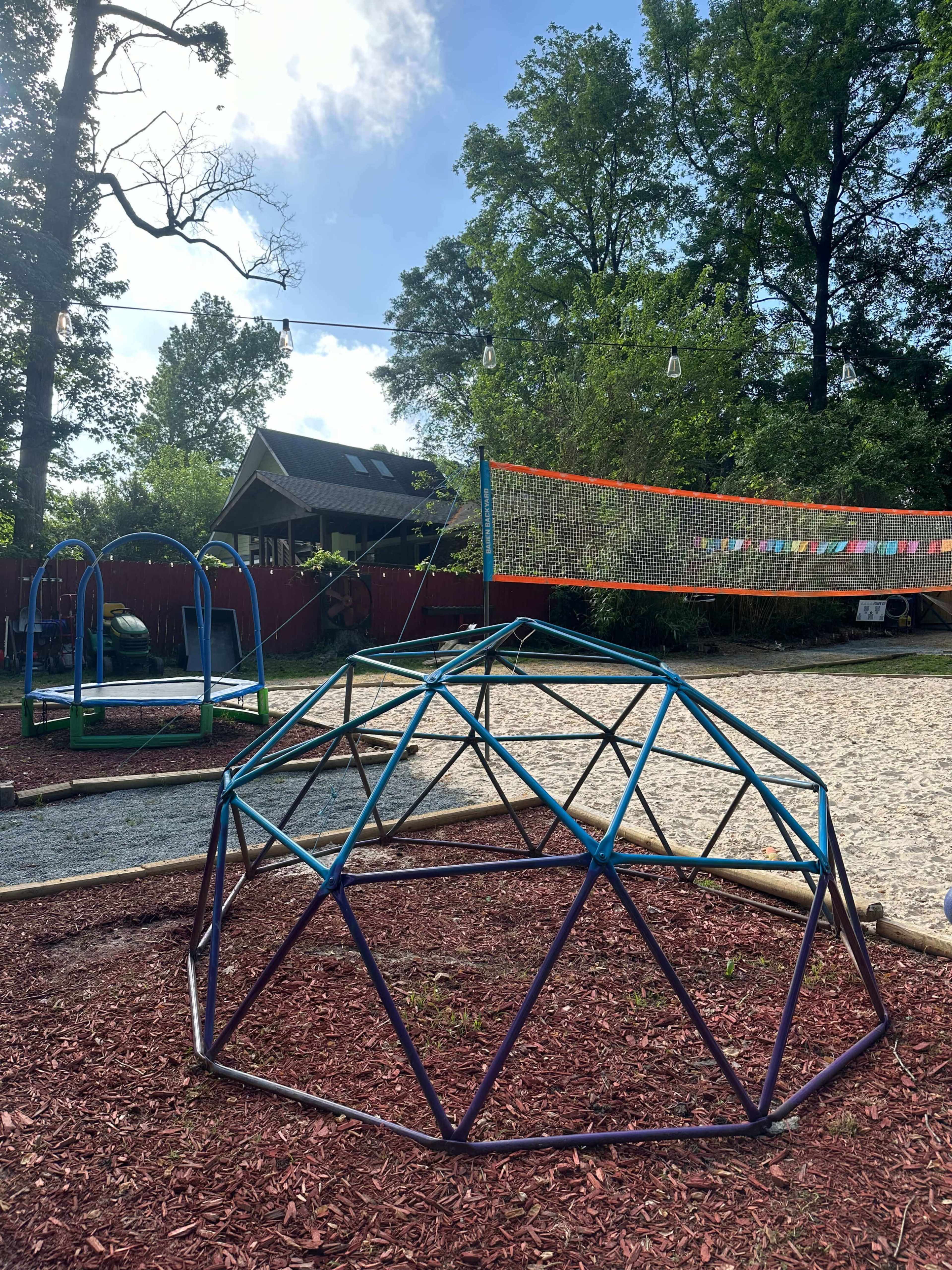 The image shows a playground area featuring a geodesic climbing dome, a volleyball net, and a shaded pavilion in the background.