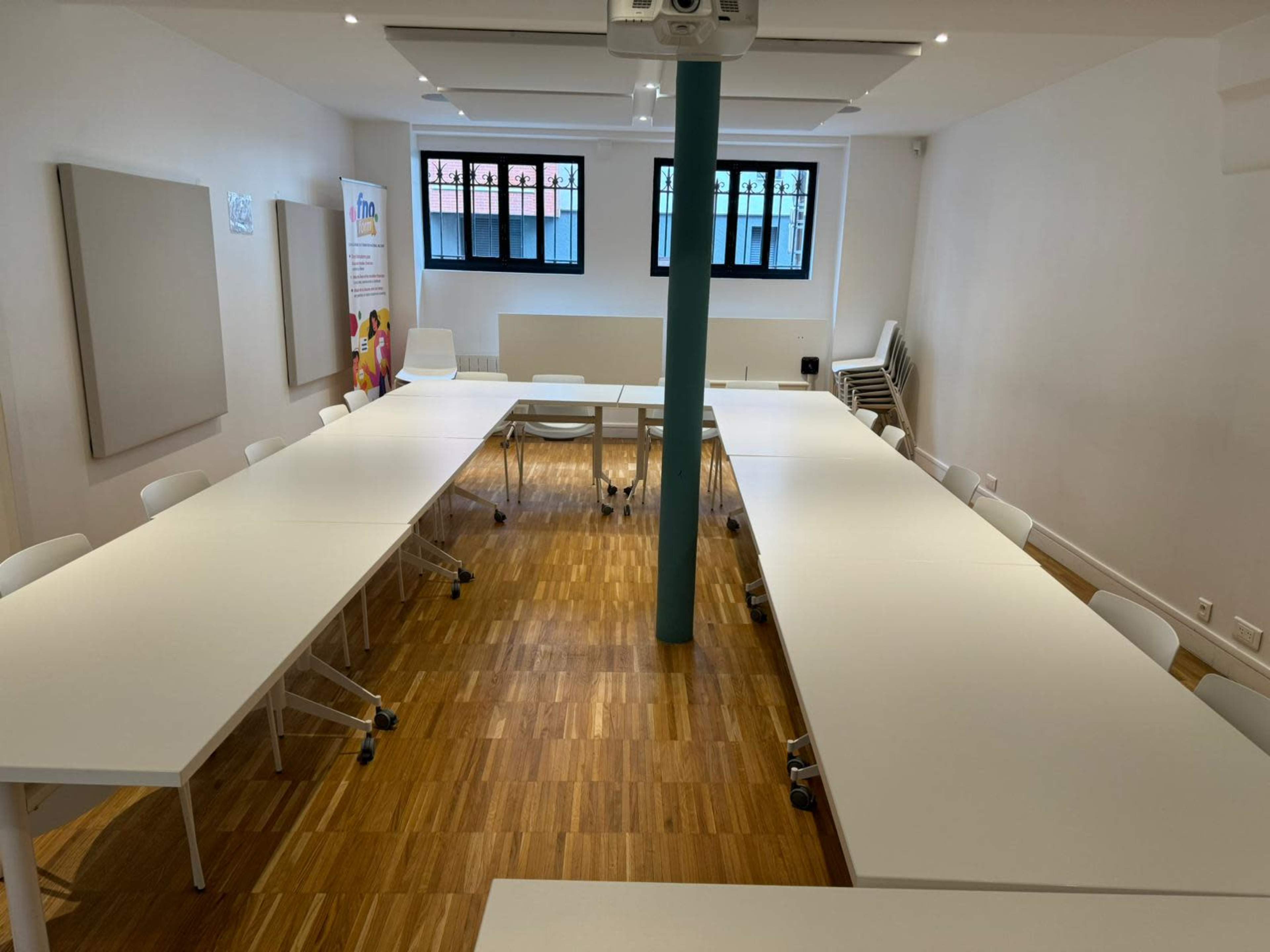 A spacious meeting room features multiple white tables arranged in a U-shape with chairs surrounding them, illuminated by overhead lighting.