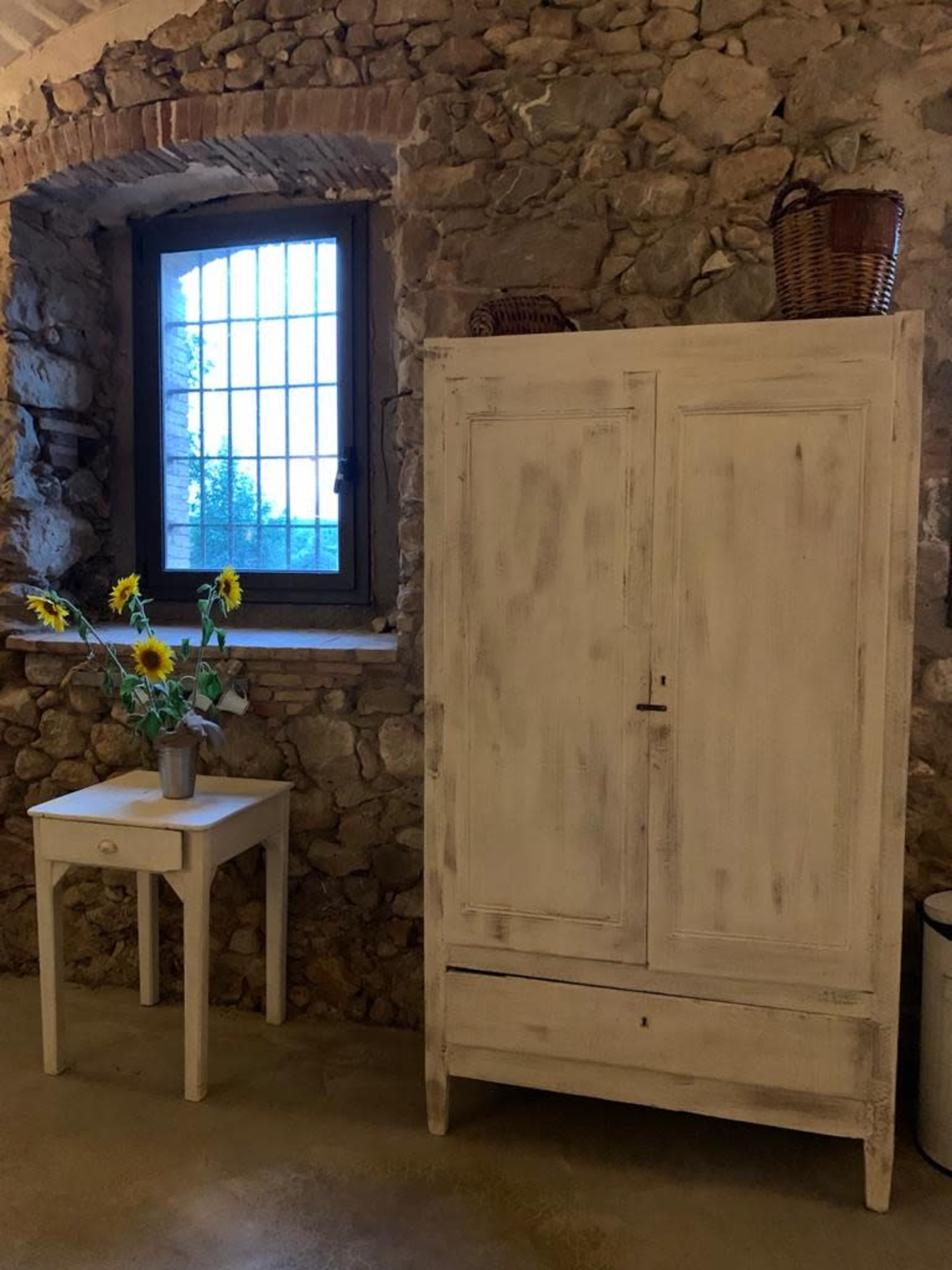 A white wooden armoire stands beside a small table with a vase of sunflowers in a room with stone walls and a window.