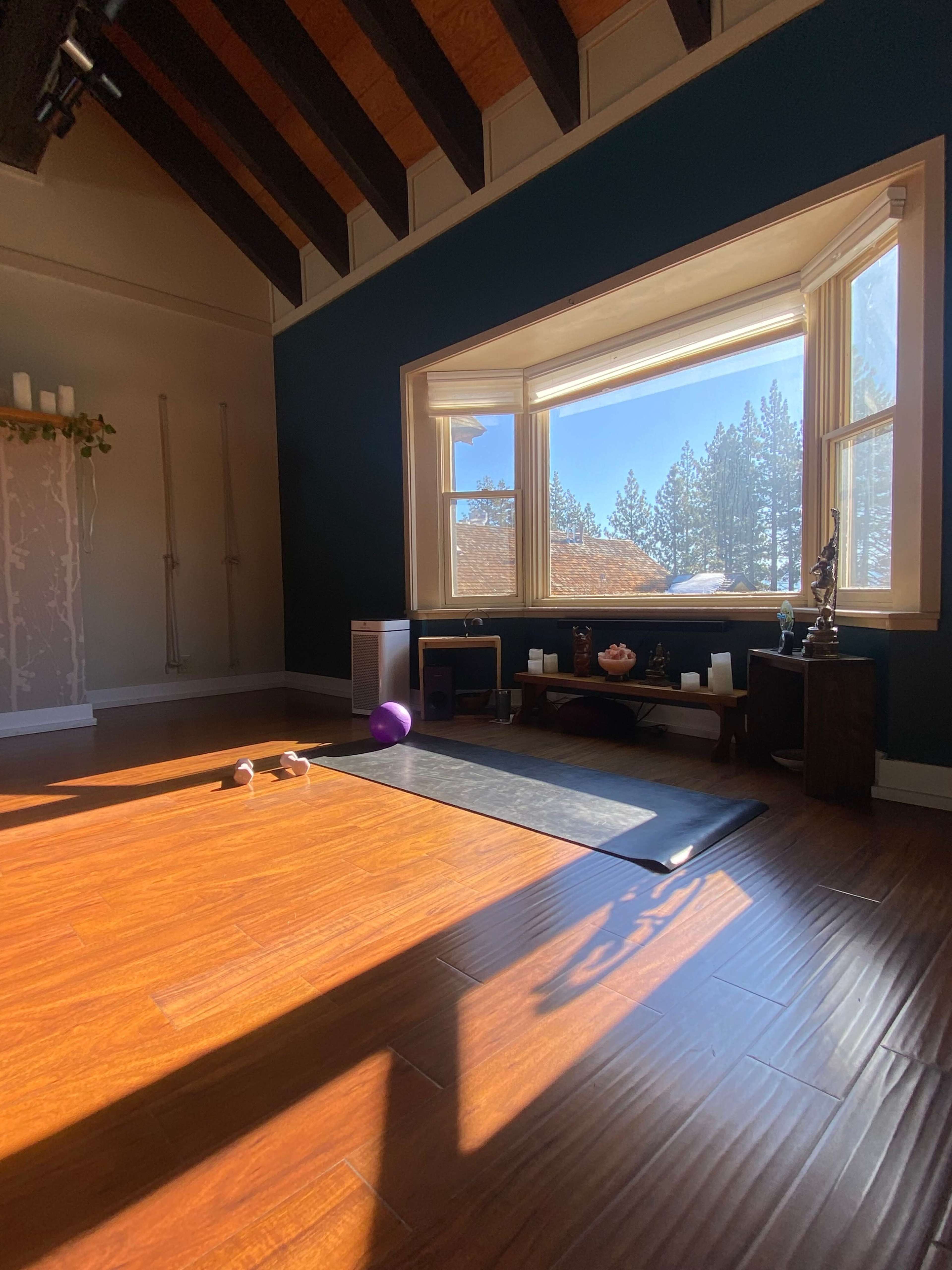 The image shows a bright, serene yoga studio with wooden floors, large windows, and minimal decor, including exercise equipment and a small altar.