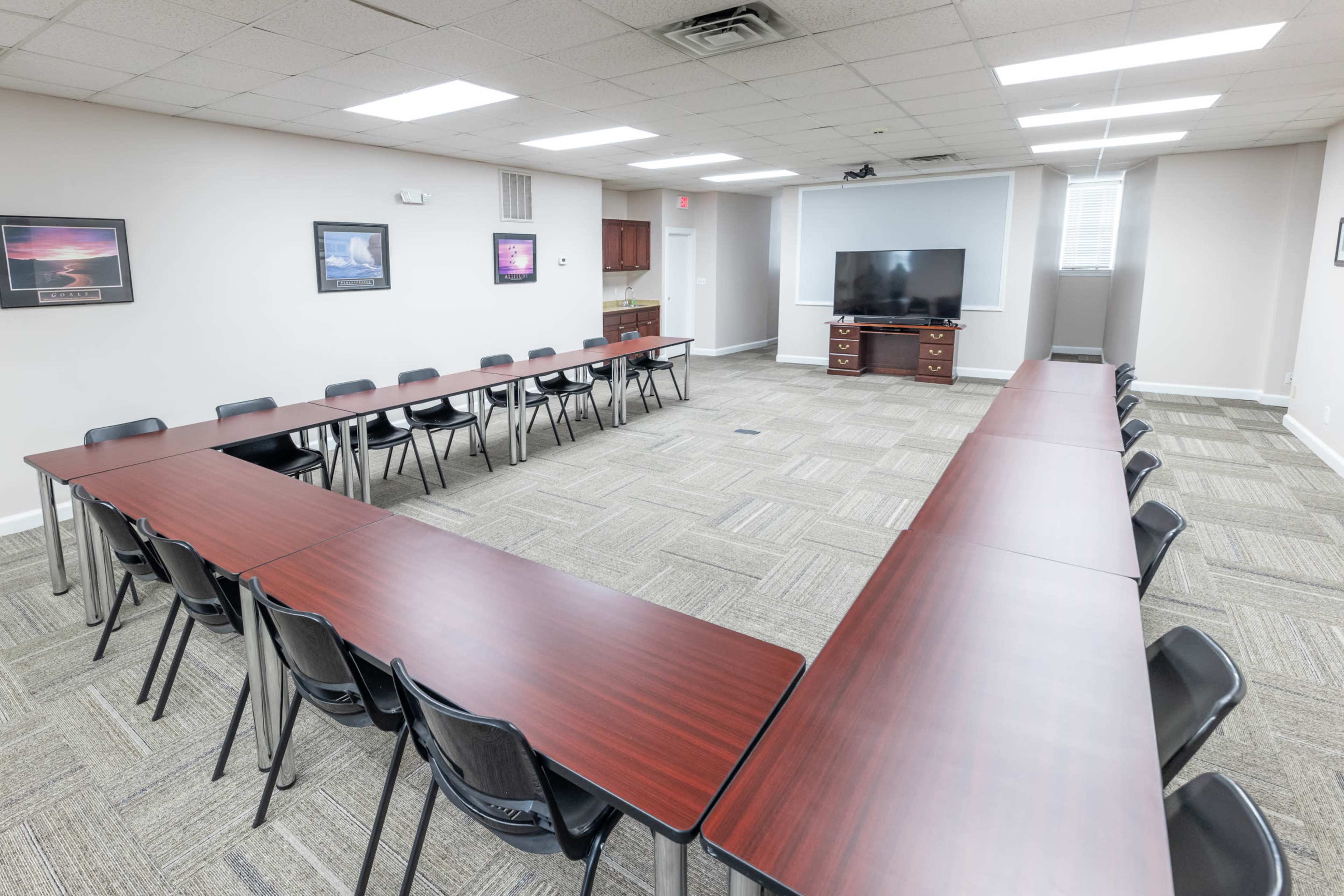 A conference room features a U-shaped arrangement of long tables and black chairs, with a large television mounted on the wall.